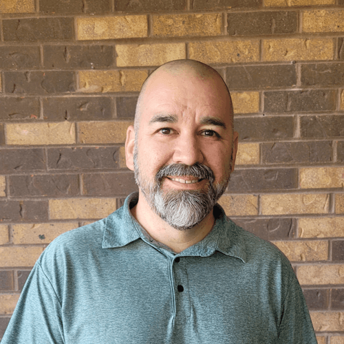 Pastor Bryan Catherman. A man with a shaved head and a gray beard smiles in front of a brick wall, wearing a teal collared shirt.