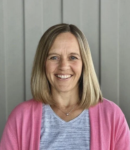 Jill Reed. A smiling woman with shoulder-length blonde hair wearing a pink cardigan and a light blue top, standing in front of a gray paneled wall.