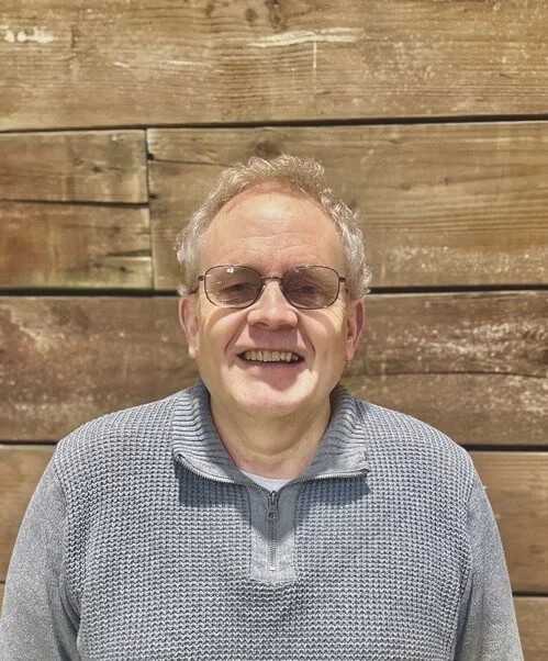 Pastor Murray Jones, smiling, with gray, curly hair, wearing glasses and a gray sweater, stands in front of a wooden wall.