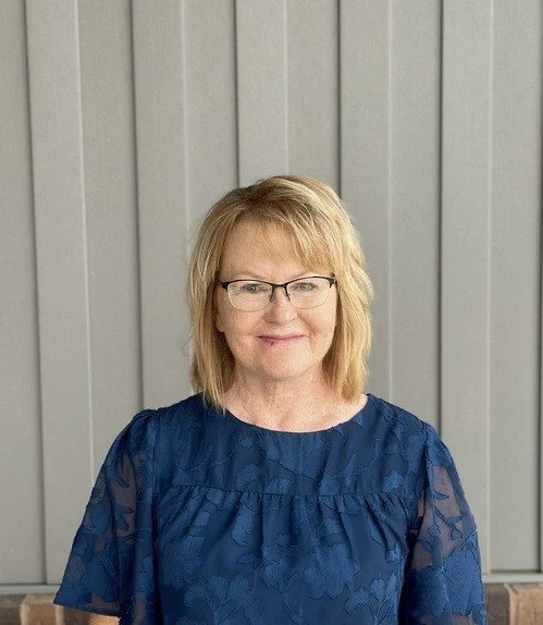 Janet Baily. A middle-aged woman with blonde hair wearing glasses and a blue blouse, standing outdoors in front of a light gray wooden wall.