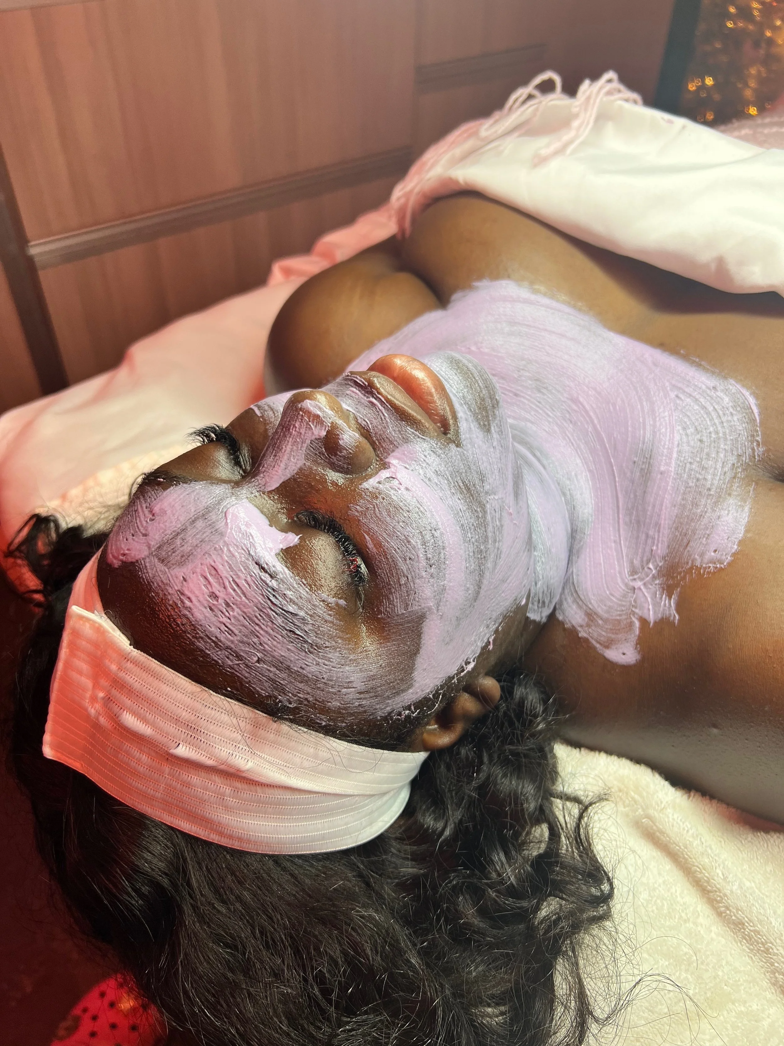 A woman with curly hair lying on a spa bed, receiving a facial treatment with a pink mask being applied to her face, with her eyes closed.