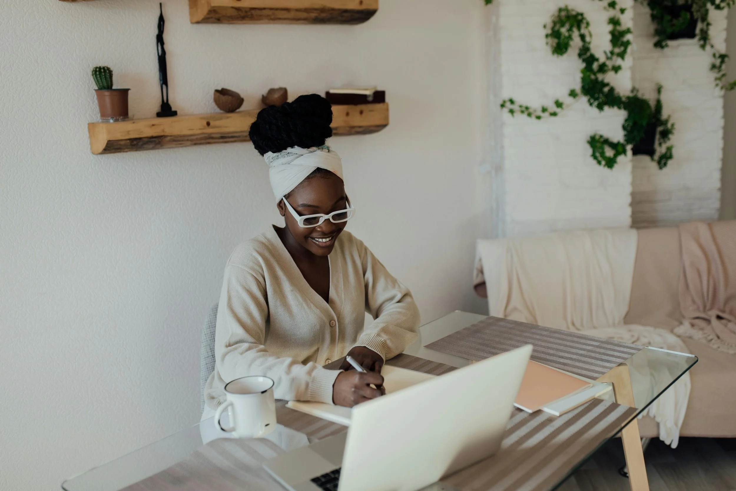 A woman with dark skin, wearing white glasses and a white headwrap, smiles while writing on a notepad at a dining table with a laptop and mug. The room is decorated with wooden shelves and green plants.