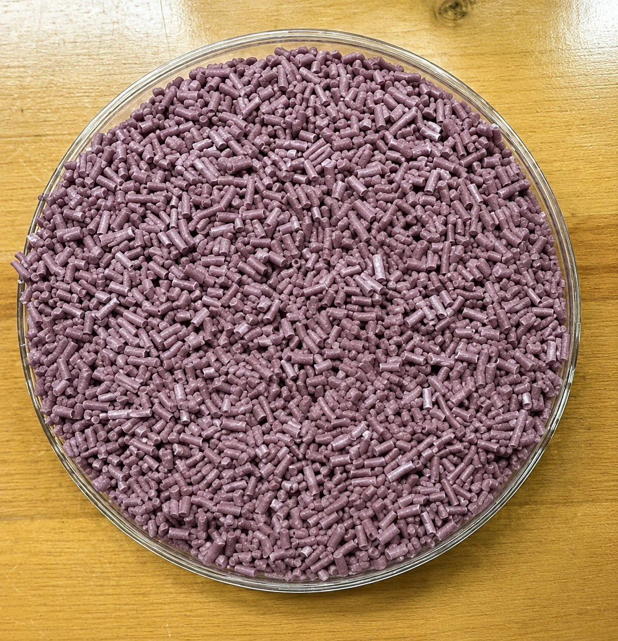 A clear round dish filled with small, purple cylindrical pellets on a wooden surface.