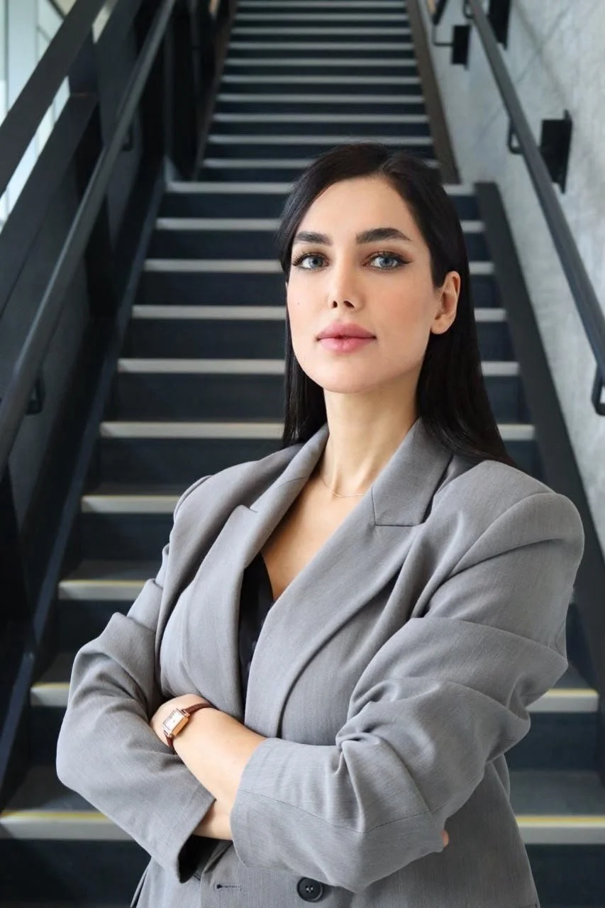A confident woman with dark hair, wearing a gray blazer, standing with arms crossed, in front of a staircase in a modern building.