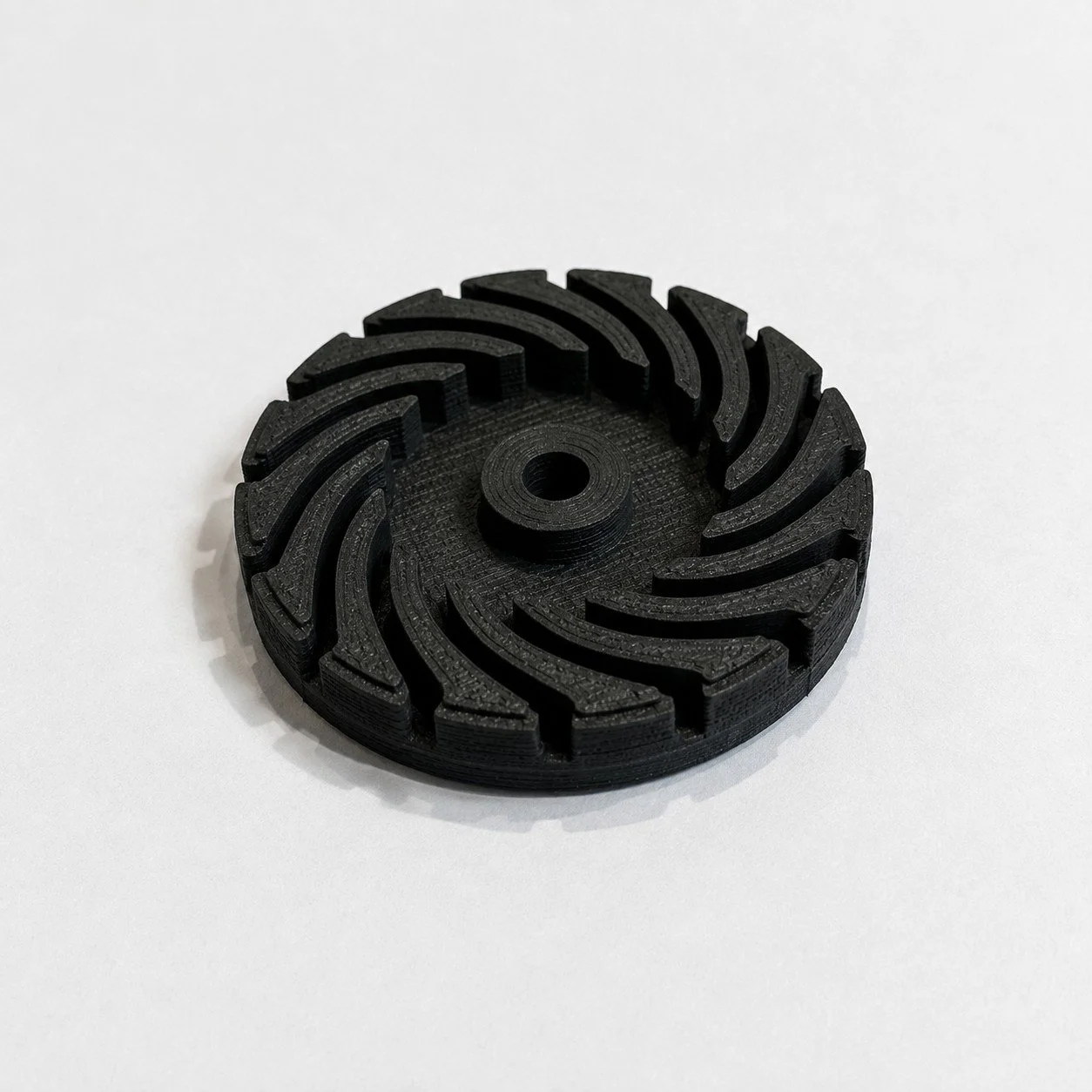 A black, circular 3D-printed object with curved grooves and a central hole, placed on a white surface.