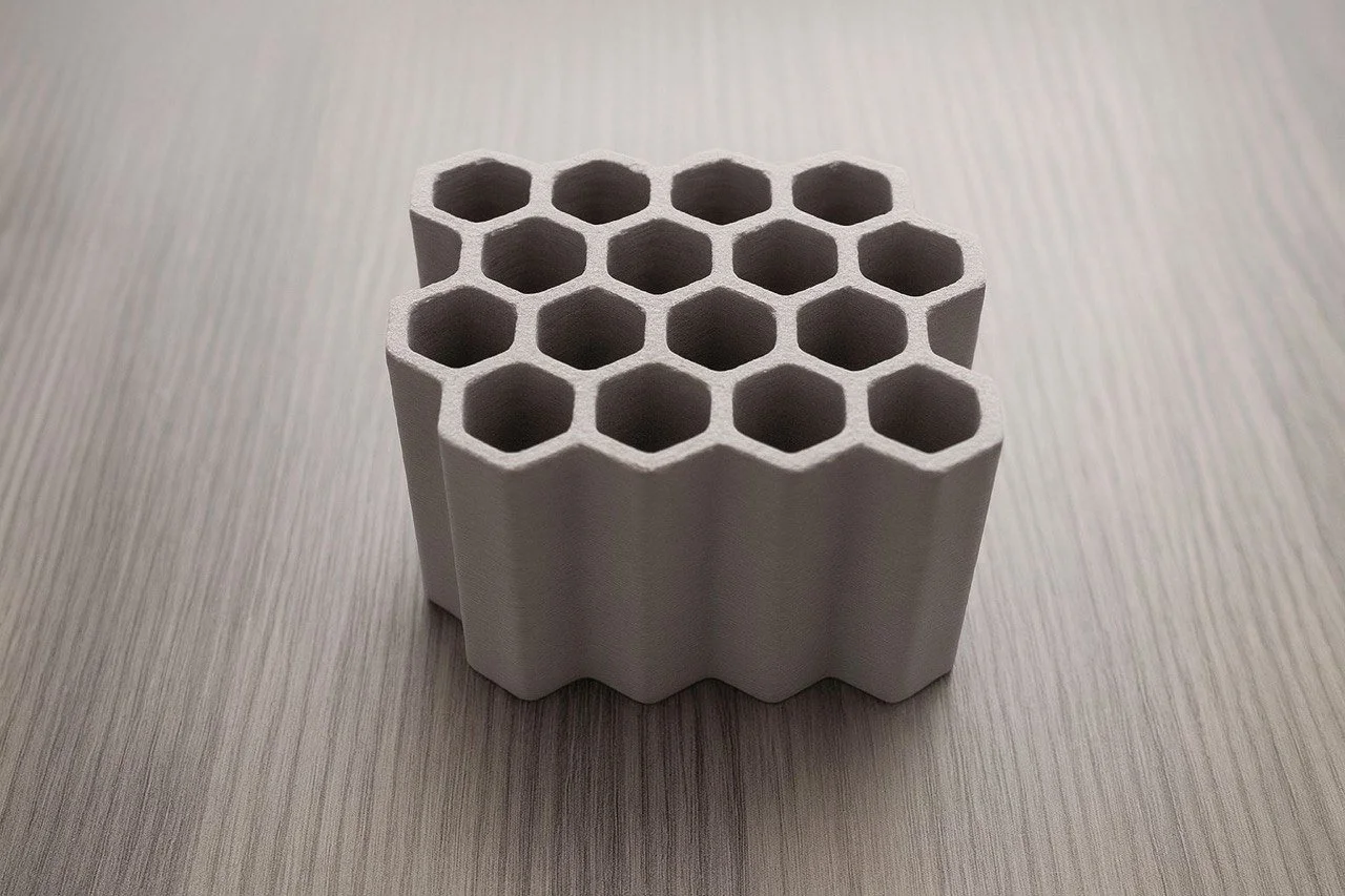 A ceramic or plastic honeycomb-shaped object with hexagonal holes, likely a filter or heat sink, placed on a light wood textured surface.