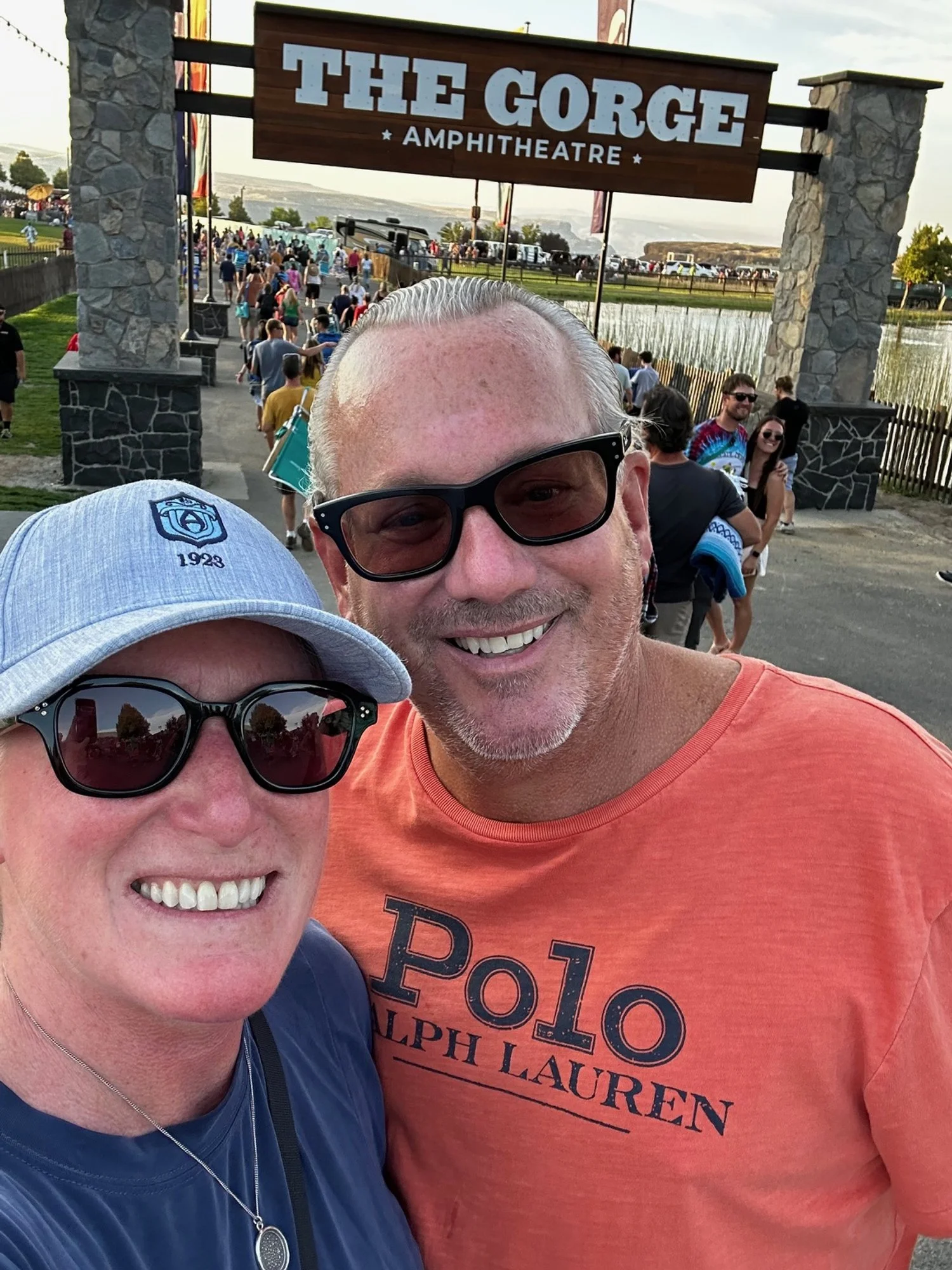Two smiling people taking a selfie in front of the entrance to The Gorge Amphitheatre, with a crowd of people in line and a lake in the background.