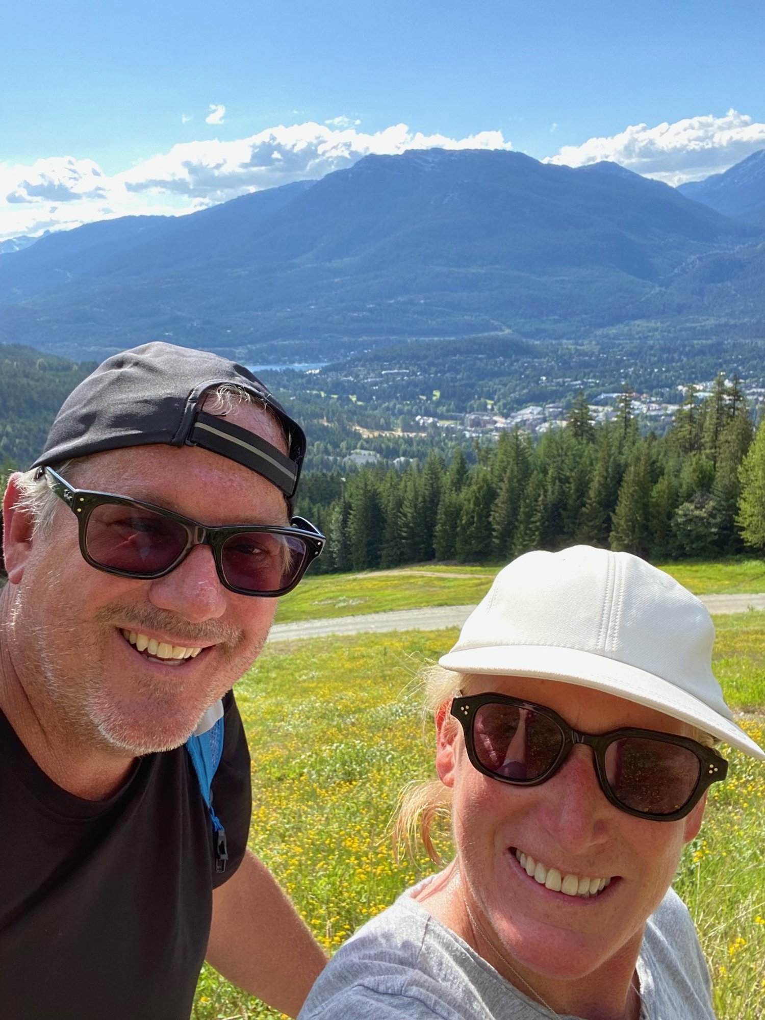 A smiling couple taking a selfie outdoors with mountains, a lake, green trees, and a field of yellow flowers in the background.