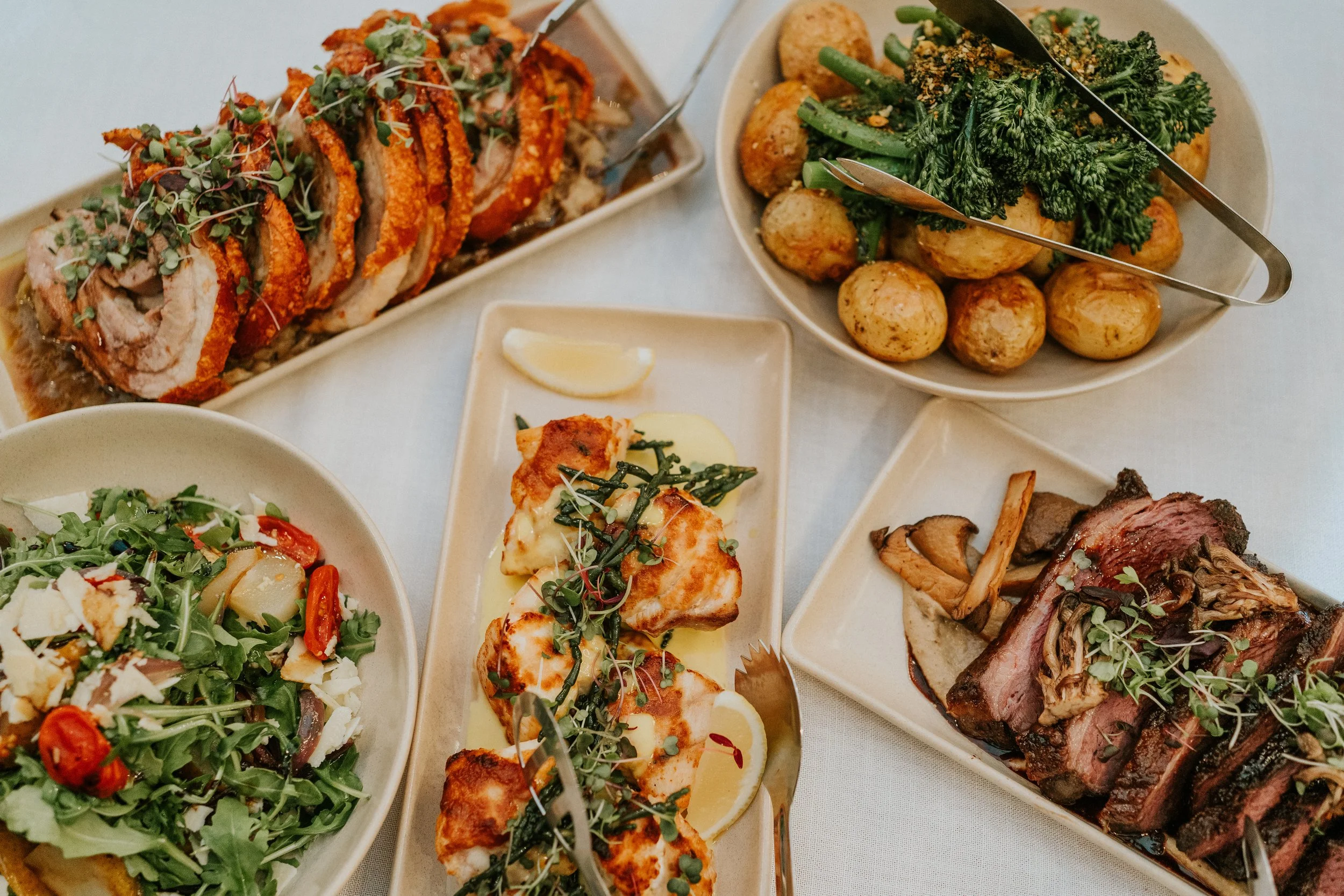 Assorted dishes on a white tablecloth including a sliced roast chicken topped with microgreens and lemon, a green salad with cherry tomatoes, a bowl of roasted potatoes and broccolini, and a plate of sliced beef with mushrooms.