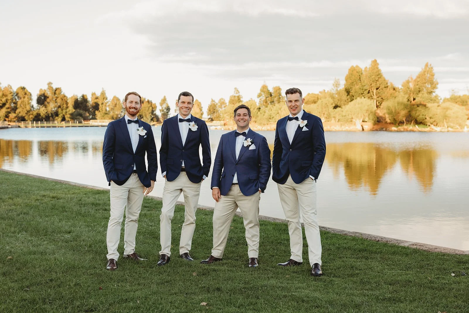 Groom and groomsmen standing by a lake during sunset, dressed in navy blazers, cream pants, and black bow ties, smiling for the camera.