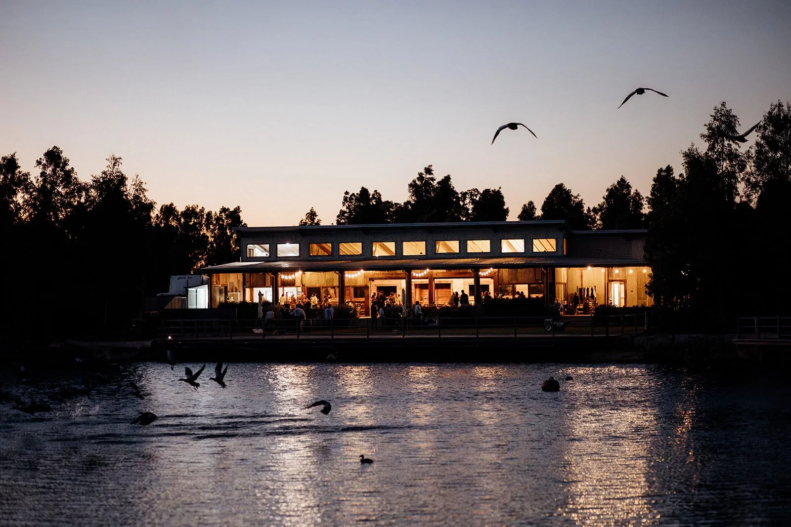 A lakeside restaurant illuminated with warm lighting at dusk, with trees and birds flying overhead and reflecting in the water.