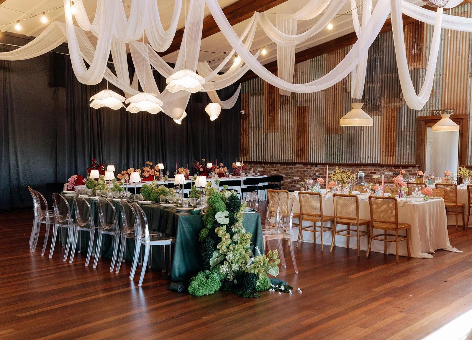 Elegant event space with decorated tables, floral arrangements, draped white fabric overhead, and pendant lights.