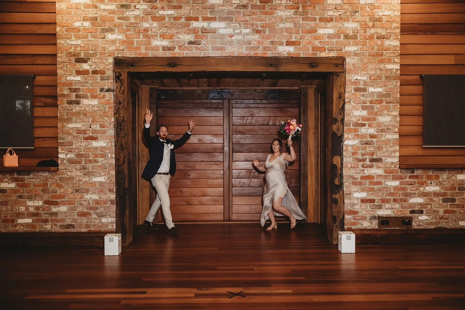 A man and woman dancing in front of a brick wall at a wedding reception, with the woman holding a bouquet of flowers.