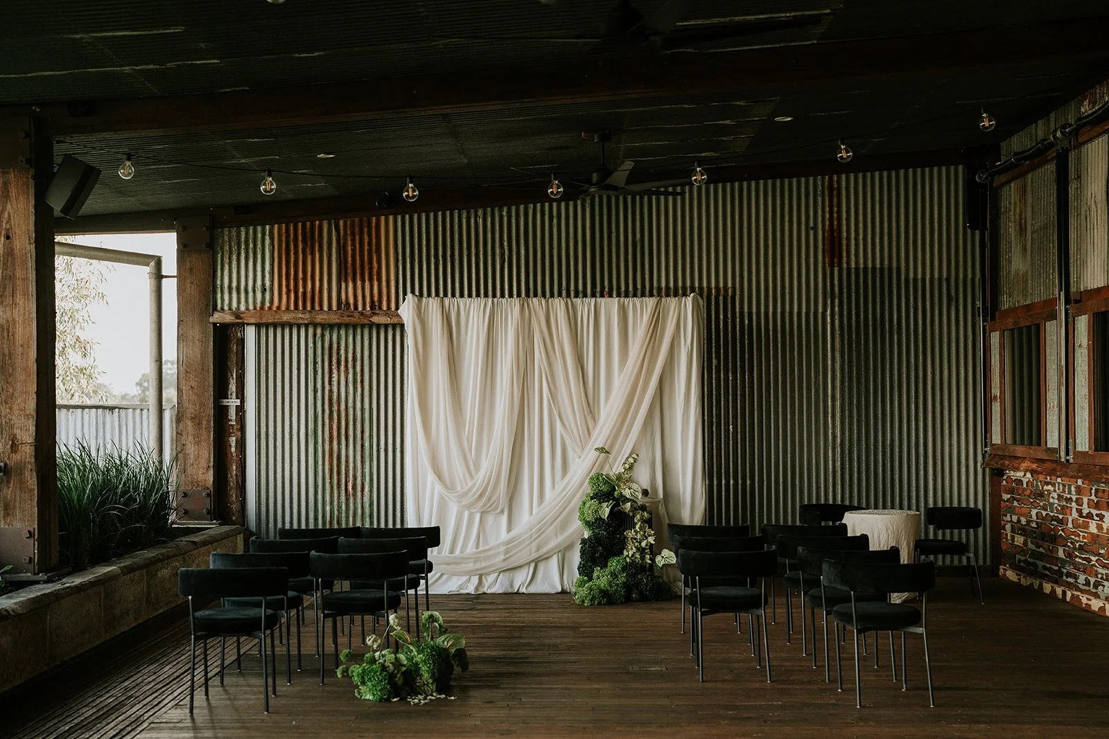 Indoor wedding ceremony setup with black chairs, white draped backdrop, and floral arrangements on a wooden floor.