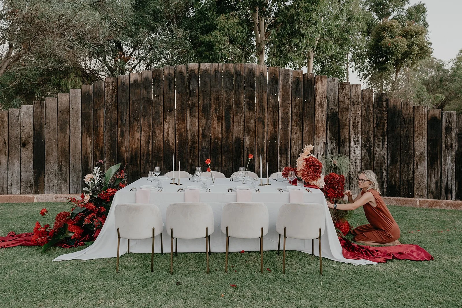 Outdoor dining setup with a long white tablecloth, beige chairs, and red floral decorations, set against a wooden fence and greenery, with a woman kneeling beside the table arranging flowers.