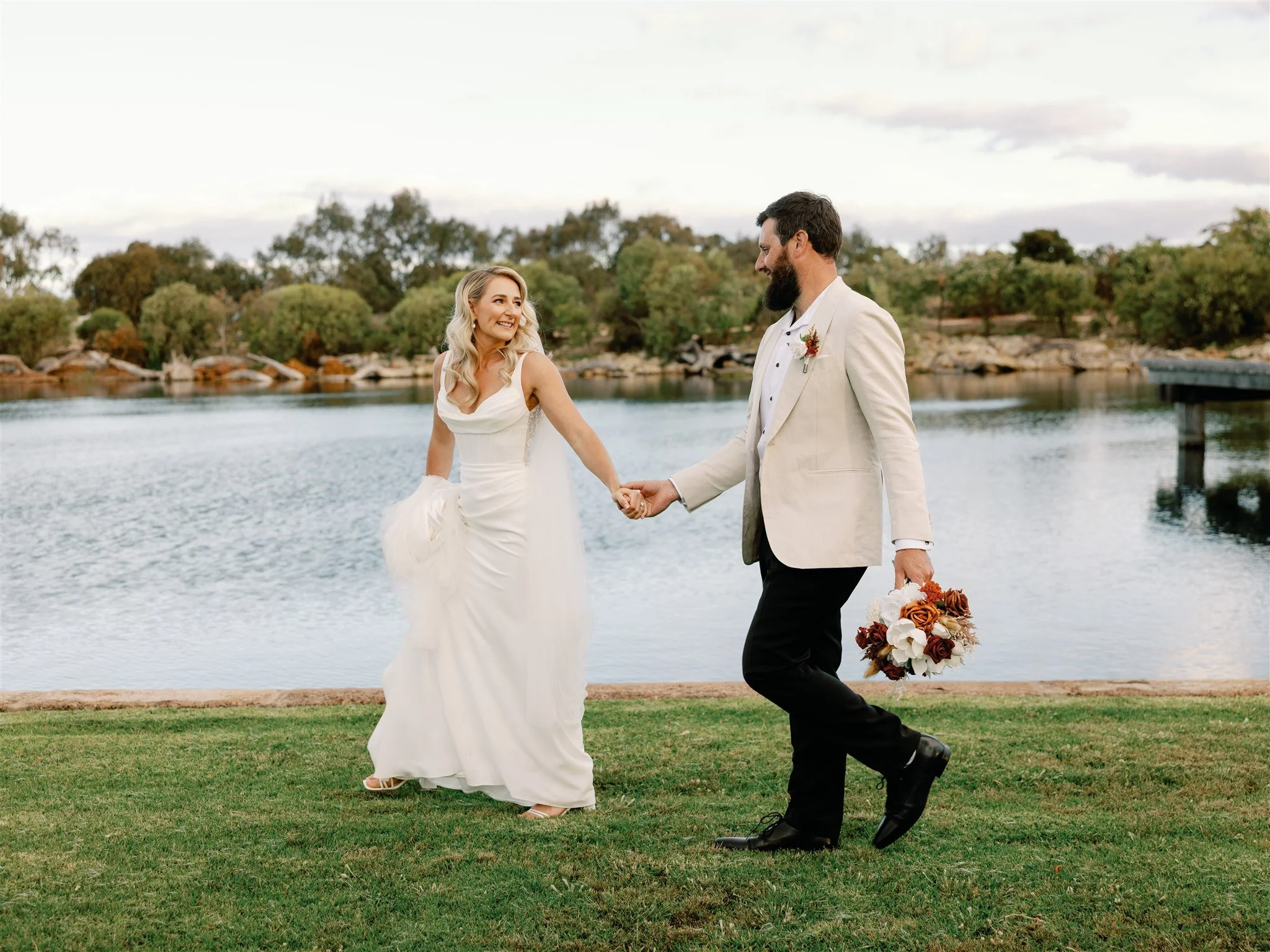 A bride and groom holding hands and smiling by a lake at sunset, with green trees and rocks in the background.