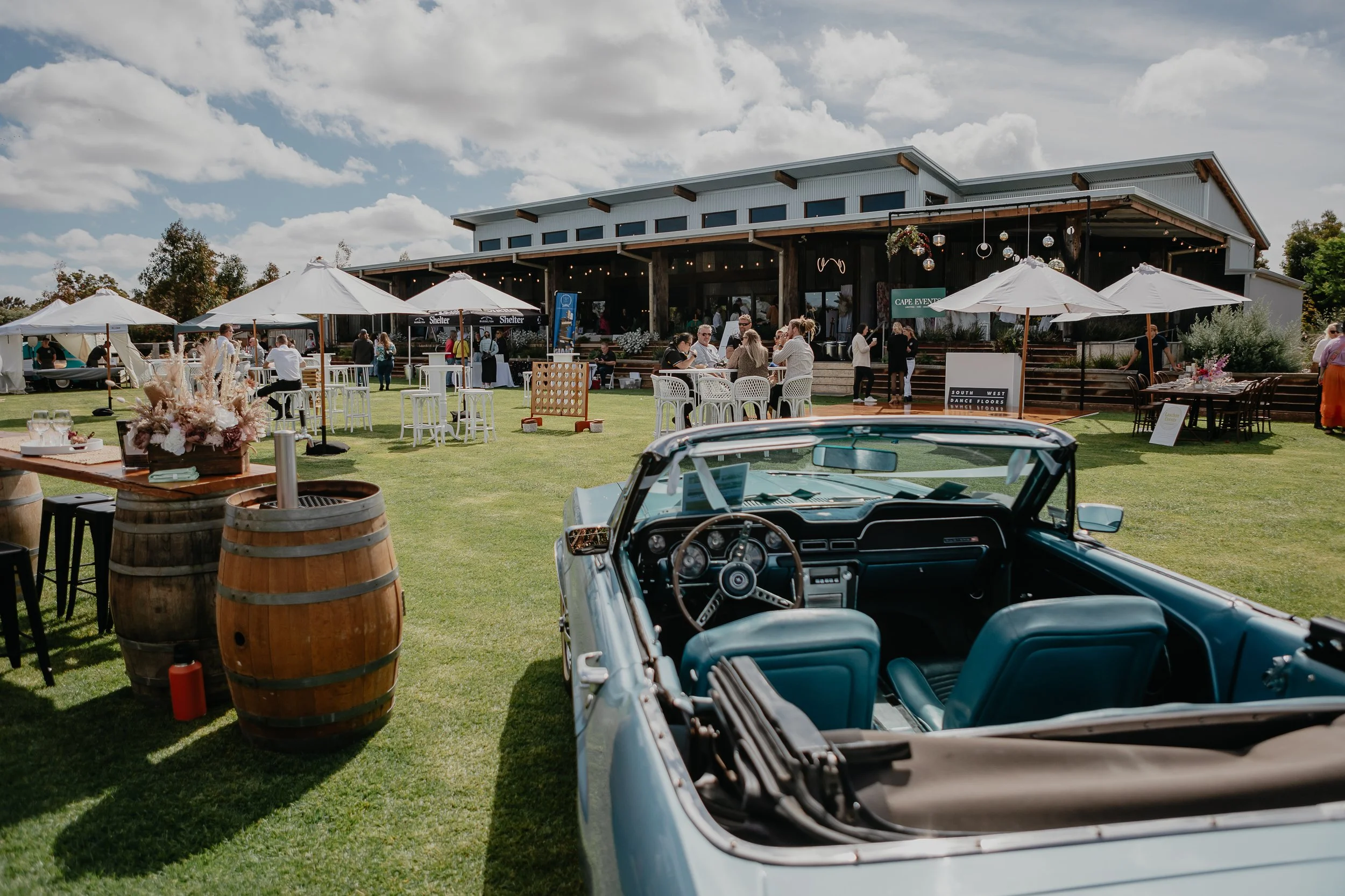 Outdoor event with people gathering on a lawn in front of a wedding and events venue, decorated with umbrellas, tables, and seating, and a vintage blue convertible car in the foreground under a partly cloudy sky.