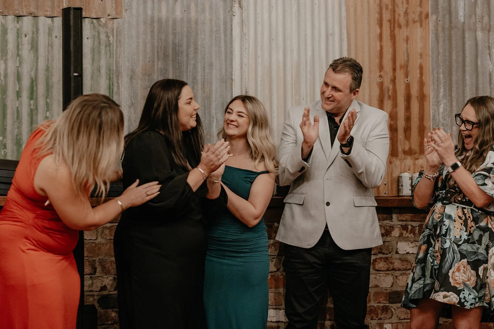 A group of five people in formal attire celebrating and clapping at an indoor event with a rustic wooden and brick wall background.