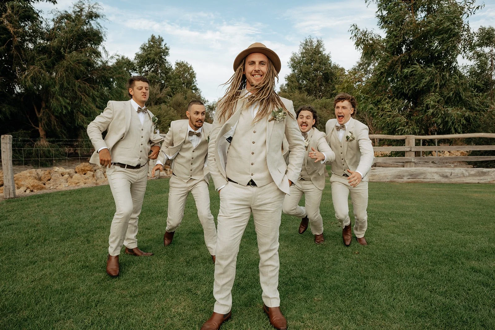 A group of six men dressed in beige suits and brown shoes, posing outdoors on a grassy area with trees in the background. One man in the front center with long hair and a hat is smiling confidently, while the other five men behind him are making playful expressions and gestures.