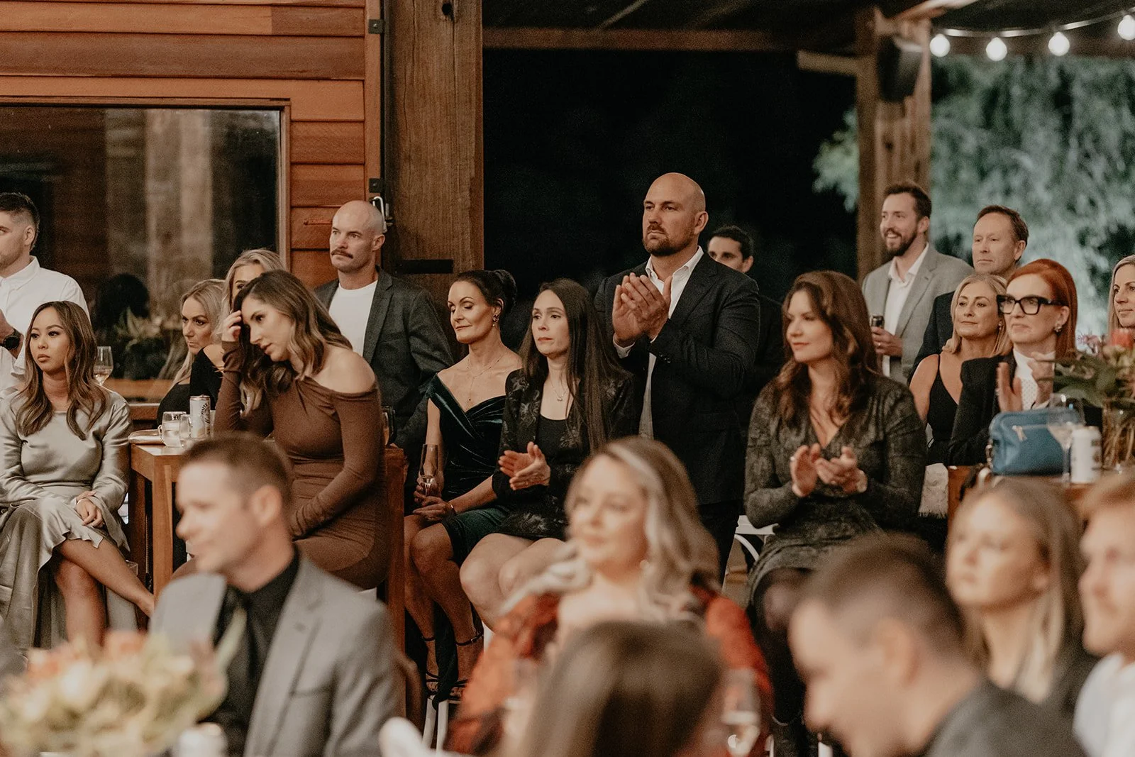 Guests at an indoor event, mostly seated, some standing, with a man in a black suit applauding. The setting has warm lighting, wooden walls, and a casual yet elegant atmosphere.