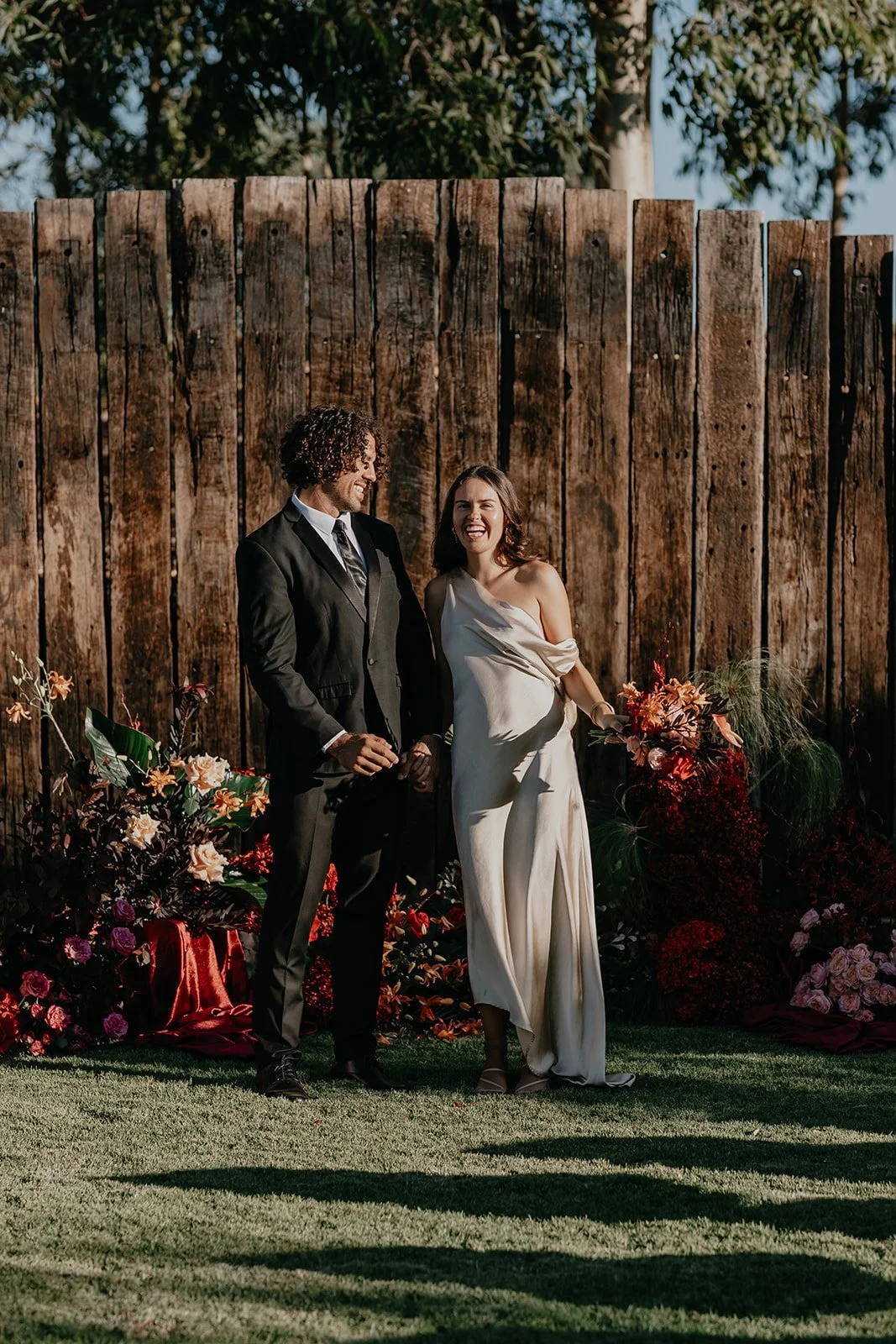 A happy couple stands outdoors with a wooden fence behind them, surrounded by vibrant flowers and greenery.