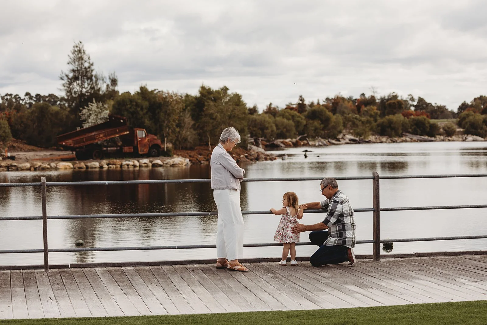 An elderly woman, a young girl, and a man are standing on a wooden deck by a lake with ducks. The woman is wearing white pants and a light-colored top, the girl is in a floral dress, and the man is kneeling in a checkered shirt. Trees and a rusty truck are visible in the background.
