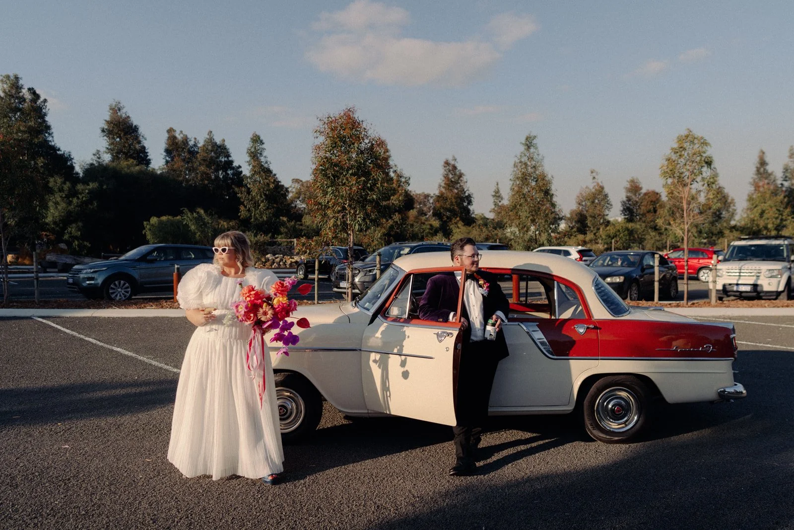 Bride in a white dress holding a large bouquet of flowers standing next to a vintage red and white car, with a groom in a dark suit holding a drink, in a parking lot with trees and parked cars in the background.
