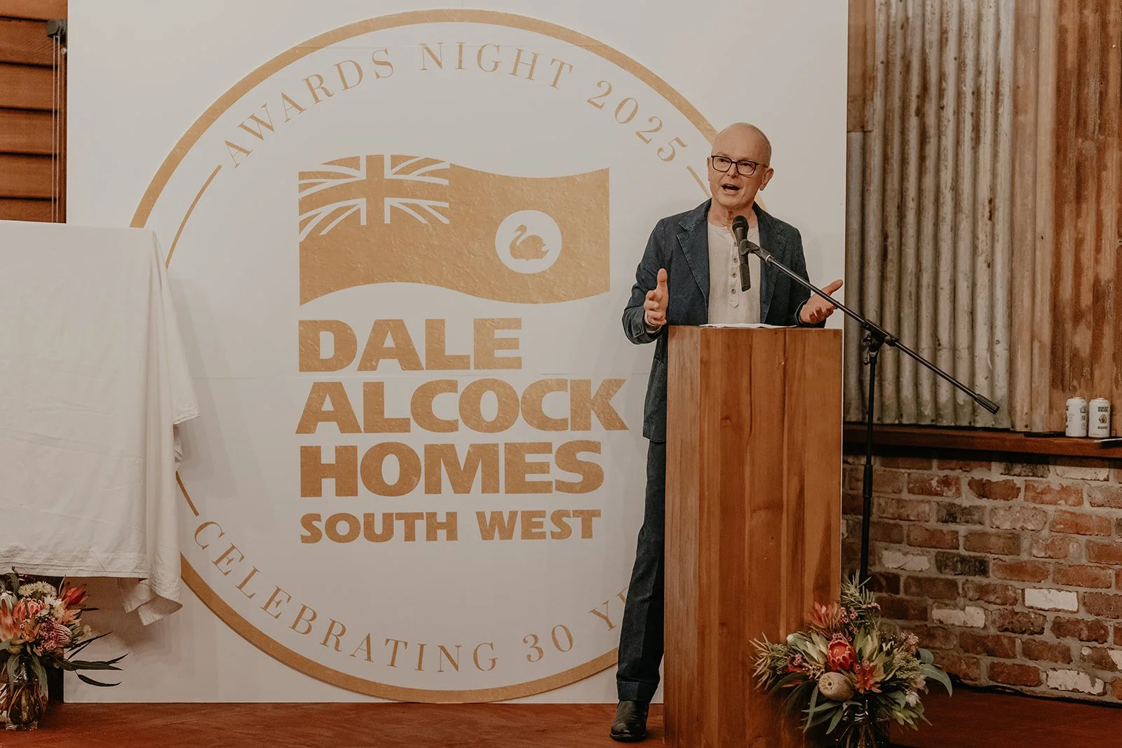 A man speaking at a podium during an awards night event with a large banner behind him that reads "Dale Alcock Homes South West," celebrating 30 years, with floral arrangements at the base of the podium.