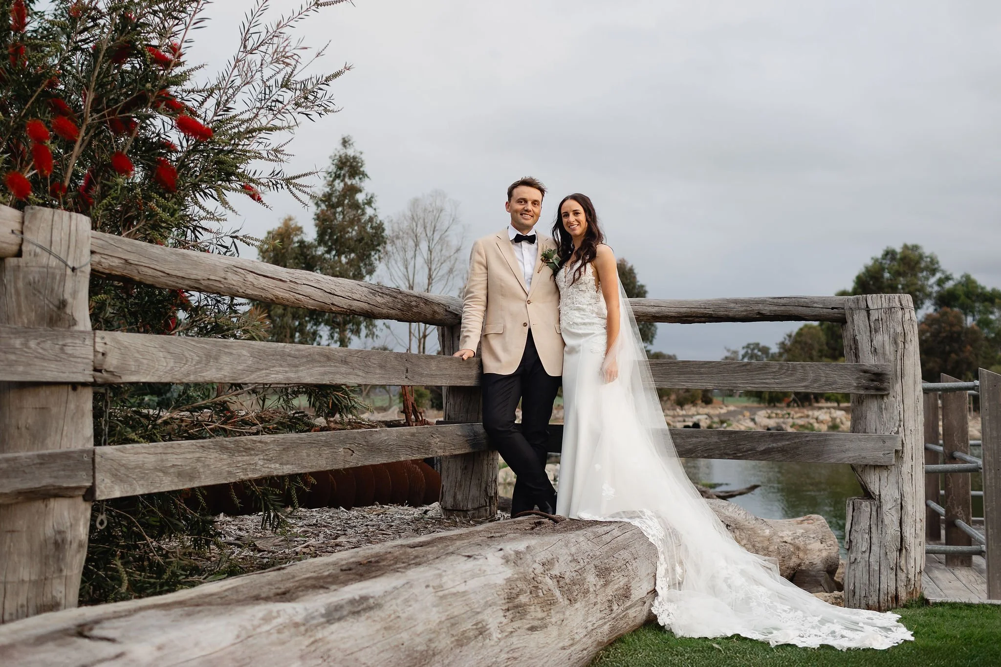 A newlywed couple standing on a rustic wooden bridge outdoors during daytime, with the bride in a white wedding dress and the groom in a beige suit, smiling.