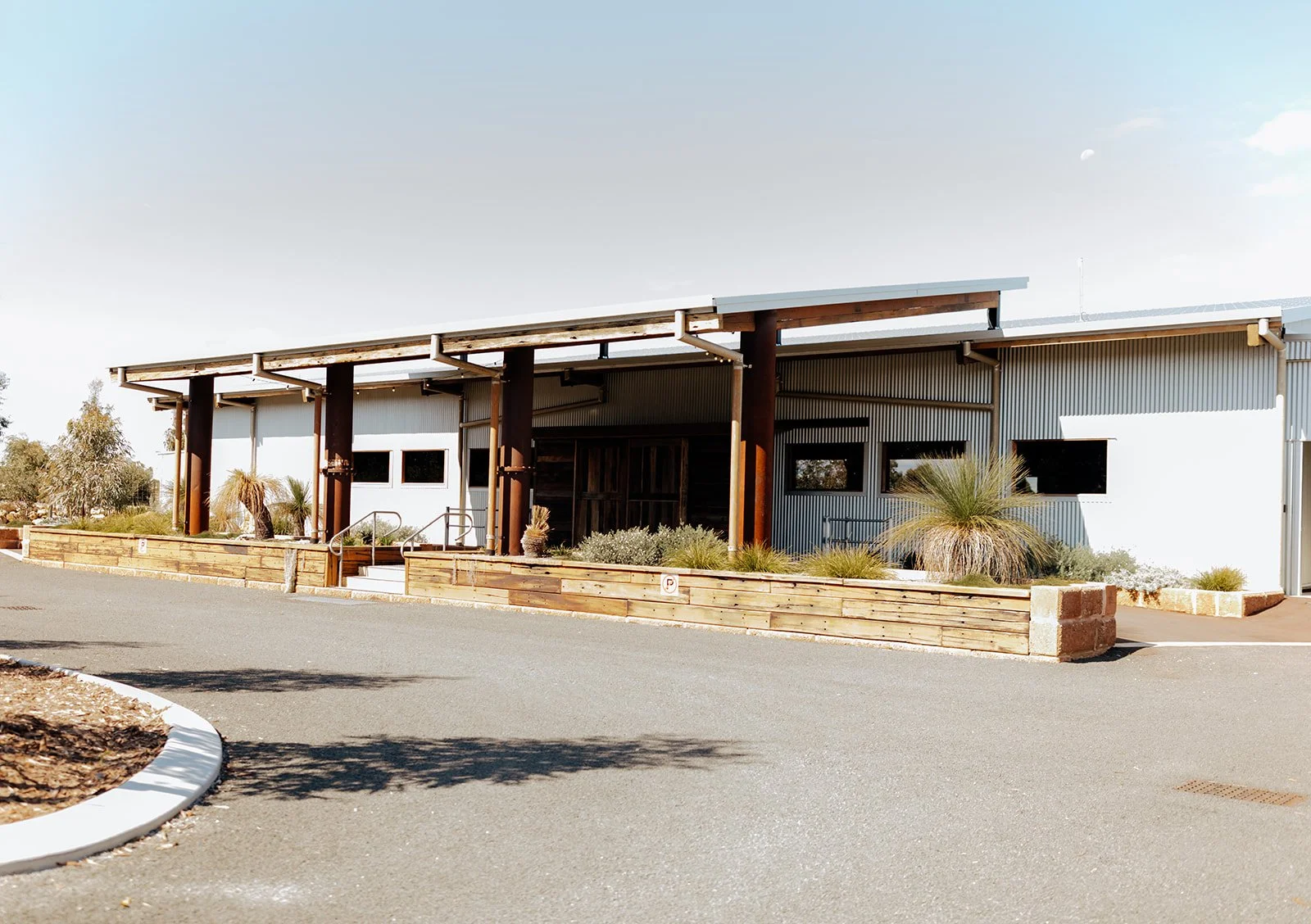 A modern industrial-style building with a metal exterior, wooden accents, and landscaped front yard featuring drought-tolerant plants, situated along a paved driveway.