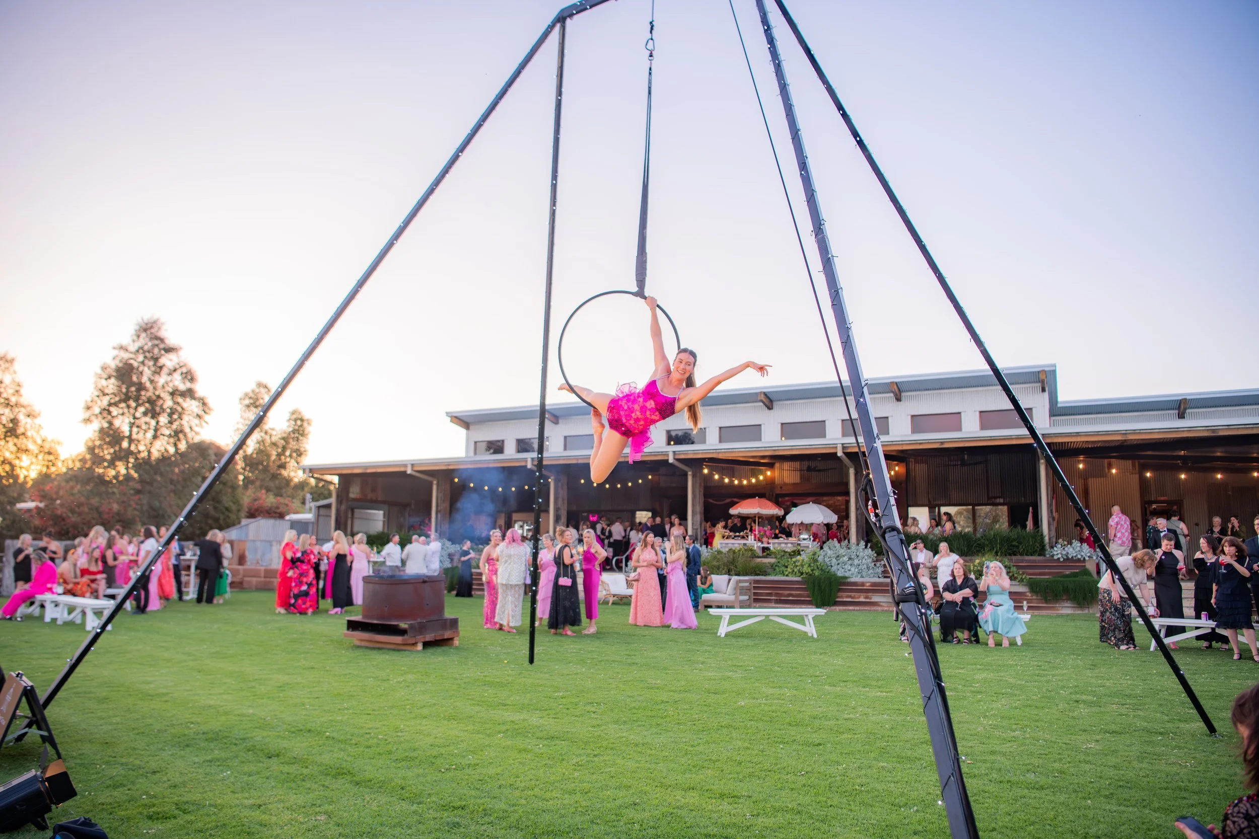 A woman performing on aerial hoop at an outdoor event in the evening, with a crowd of people in formal attire in the background.