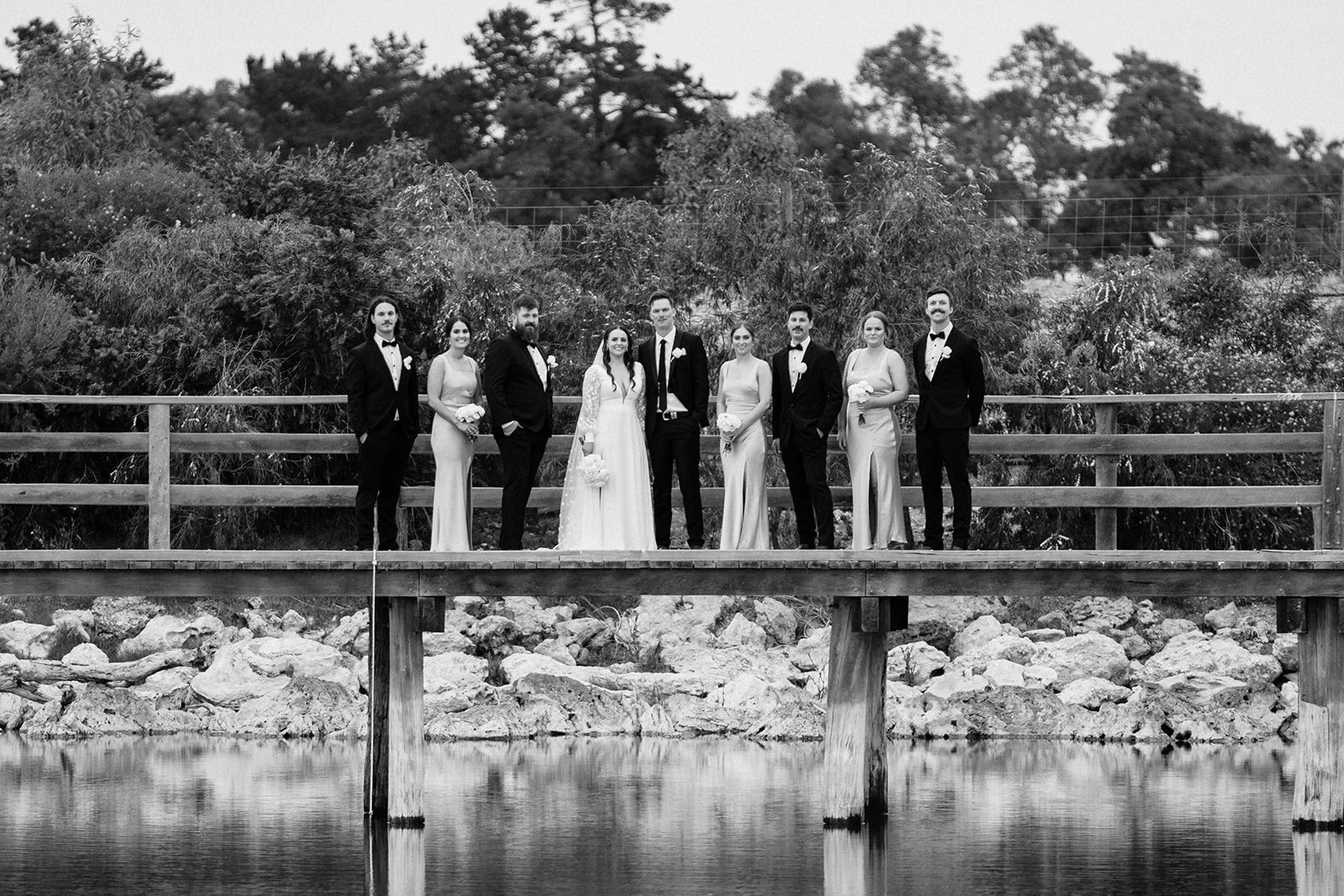 Black and white photo of ten people dressed in formal wedding attire standing on a wooden bridge over water, with trees and bushes in the background.