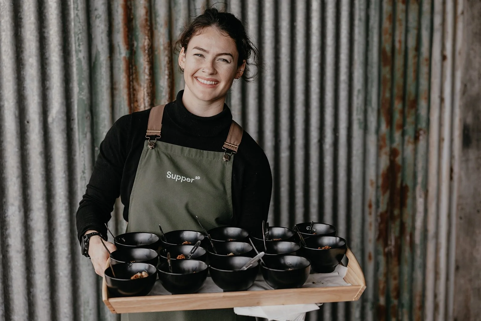 A woman smiling and holding a tray with black bowls of food in front of a corrugated metal wall.