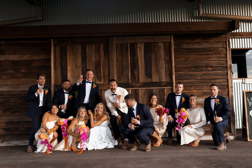 A group of eleven people, including the bride and groom, dressed in wedding attire, are posing for a photo in front of a wooden structure. They are holding flowers and drinks, smiling and making celebratory gestures.