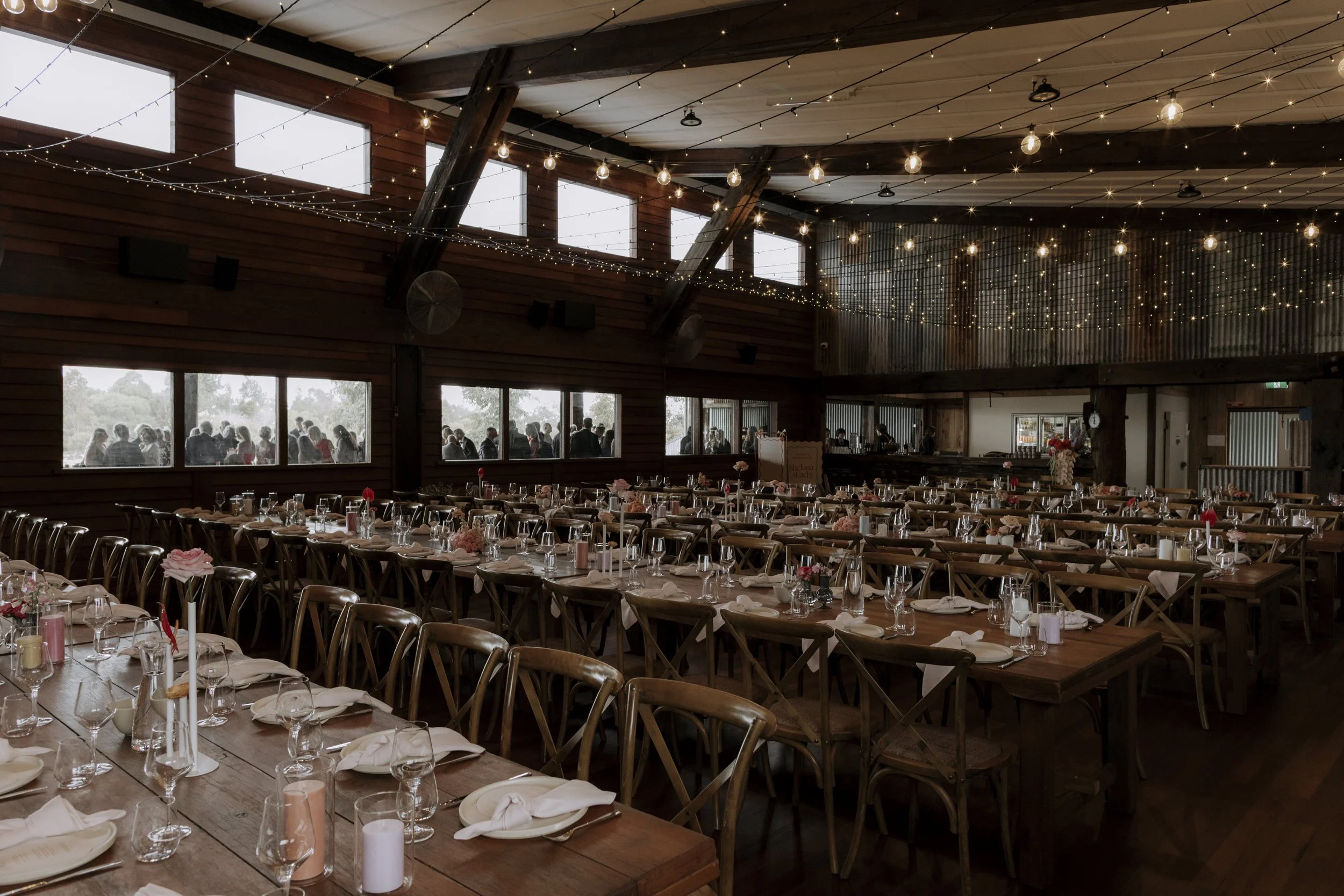 Interior of a rustic event hall set up for a celebration with long wooden tables, elegant table settings, and string lights hanging from the ceiling.