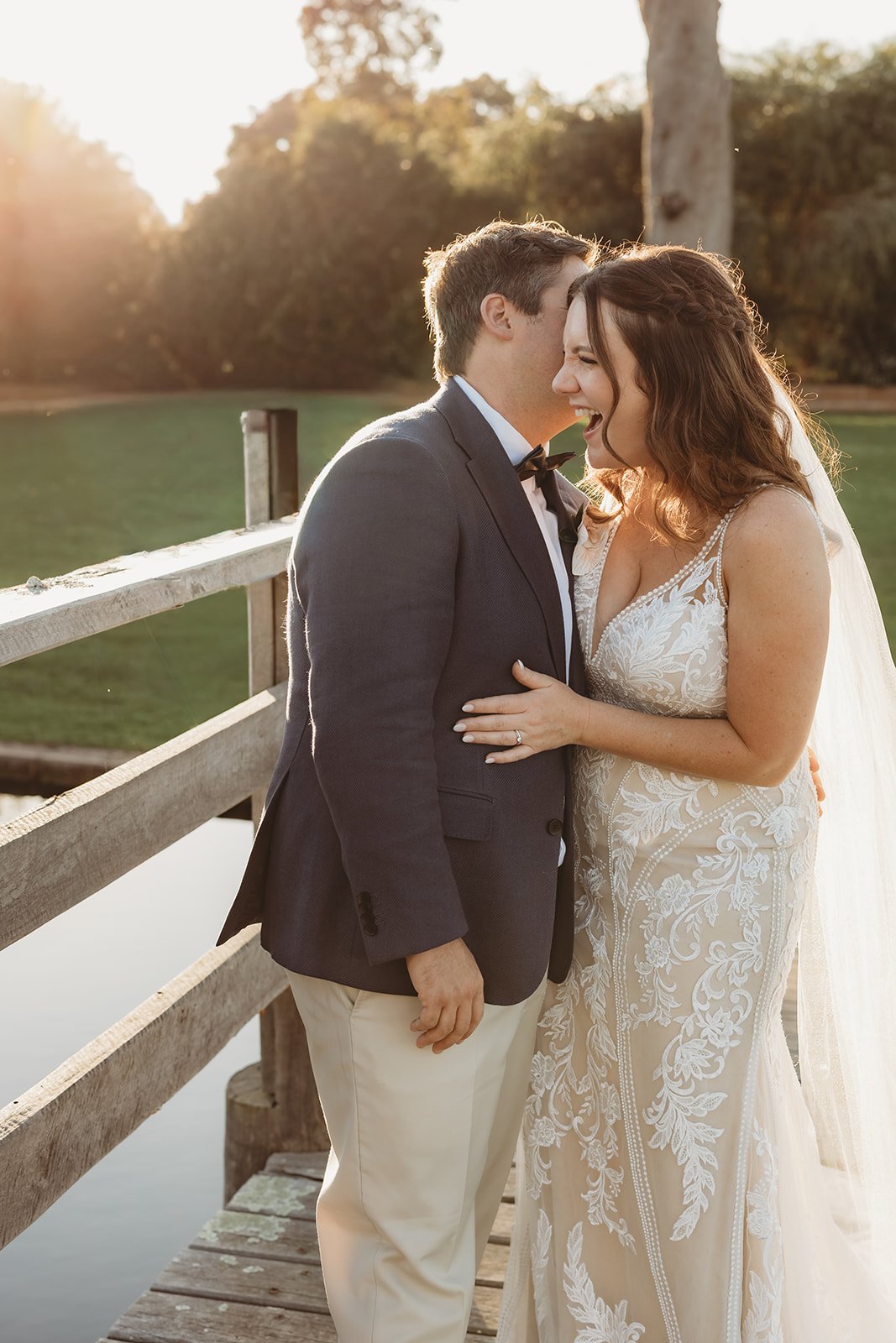 A bride and groom sharing a joyful moment on a wooden dock near a body of water during sunset.