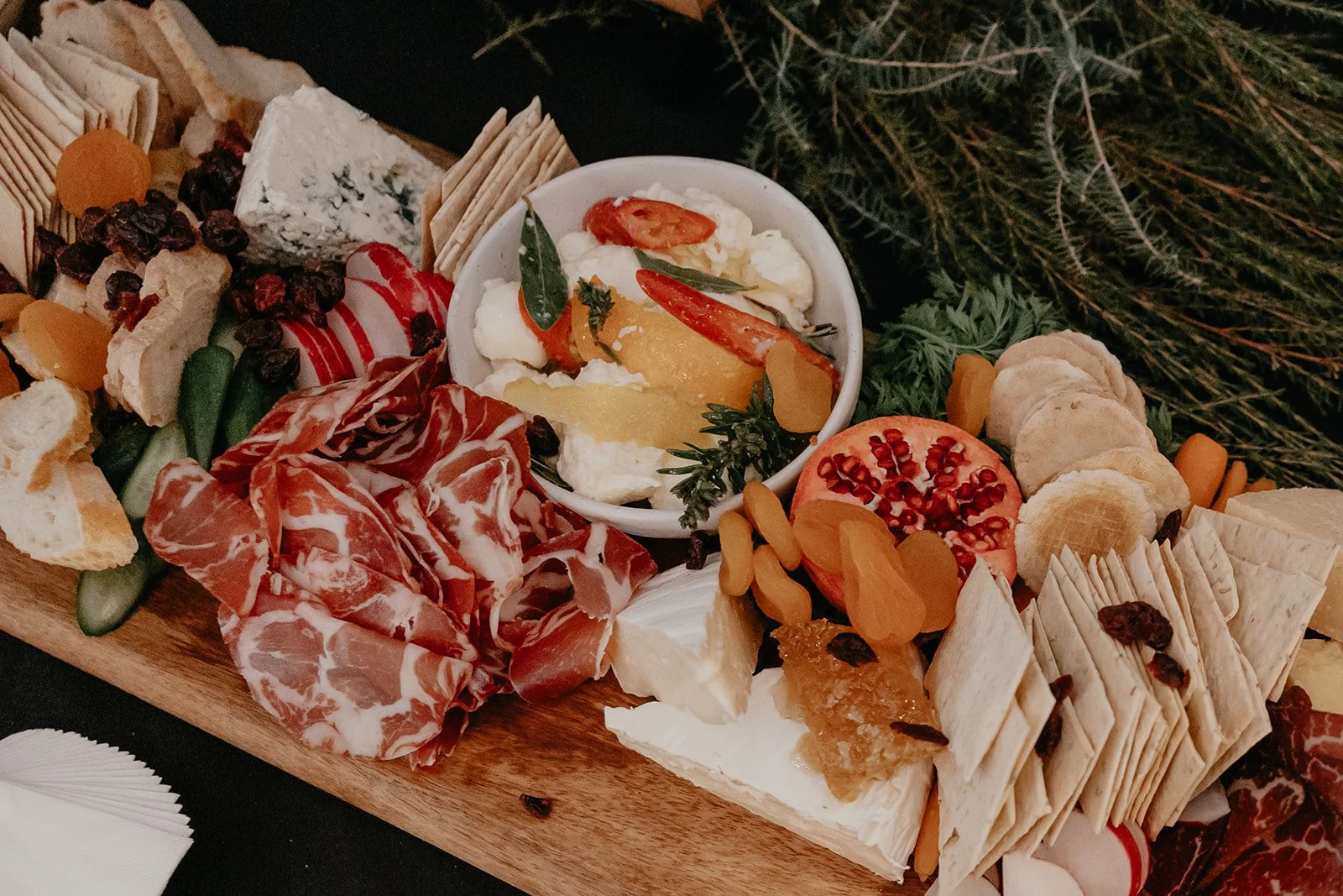A cheese and charcuterie board with sliced meats, cheeses, crackers, dried fruits, fresh tomato, and garnishes, served on a wooden platter with greenery in the background.