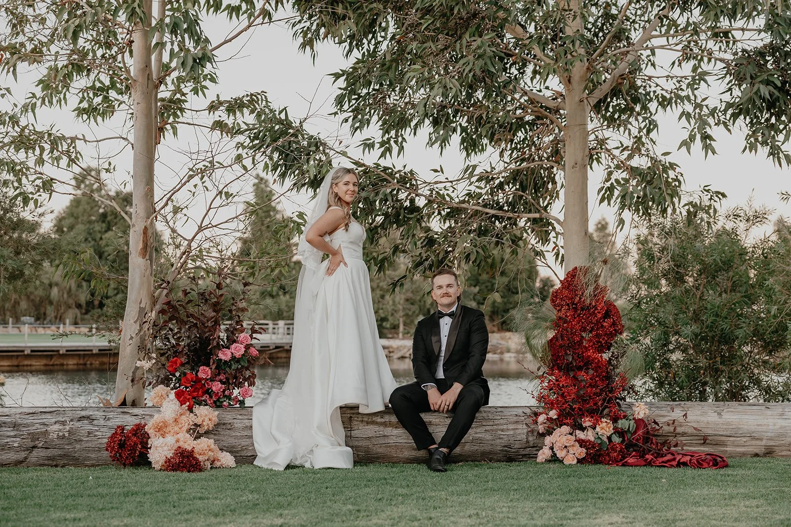 A bride standing with her hand on her hip and a groom sitting on a log under trees, surrounded by flowers, by a body of water during a wedding photoshoot.