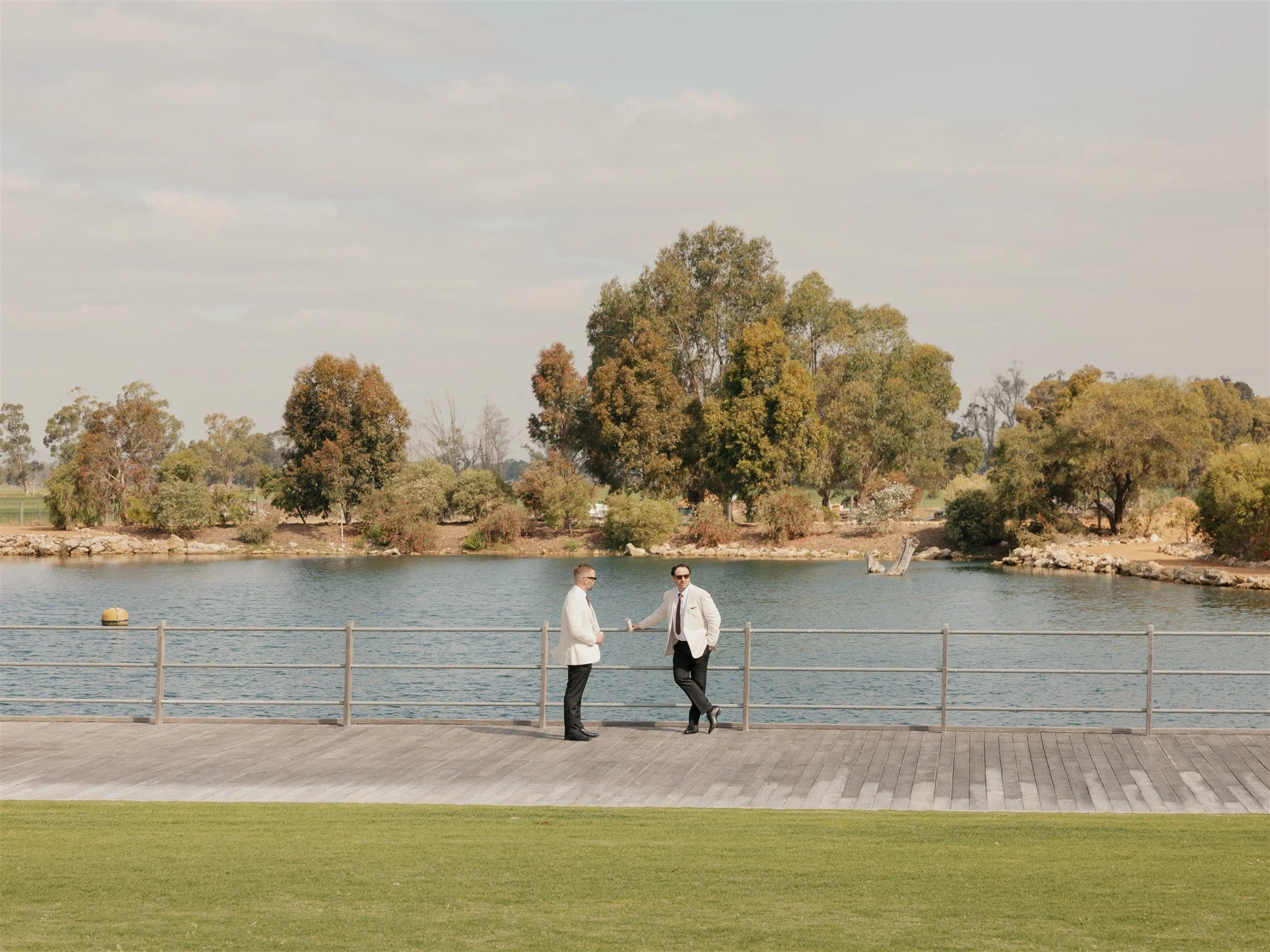 Two men in suits standing and chatting on a wooden dock by the water, with trees and a partly cloudy sky in the background.
