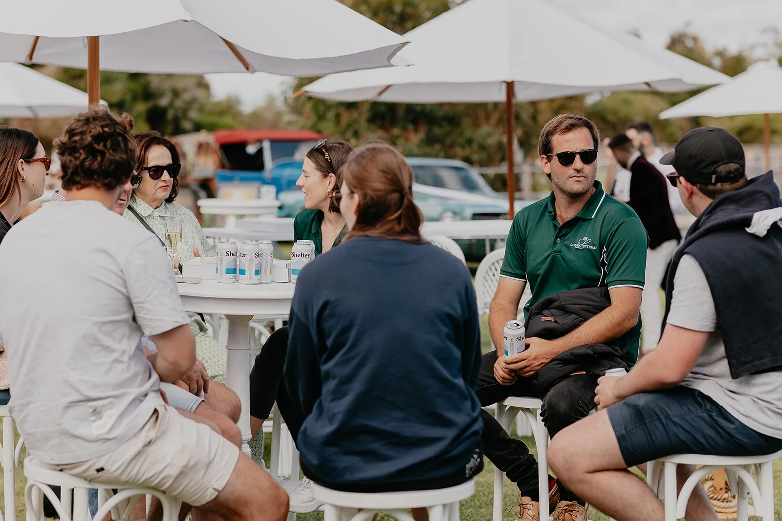 Group of people sitting around a table outdoors, chatting and holding cans of Shelter beer, with umbrellas overhead and vintage cars in the background.
