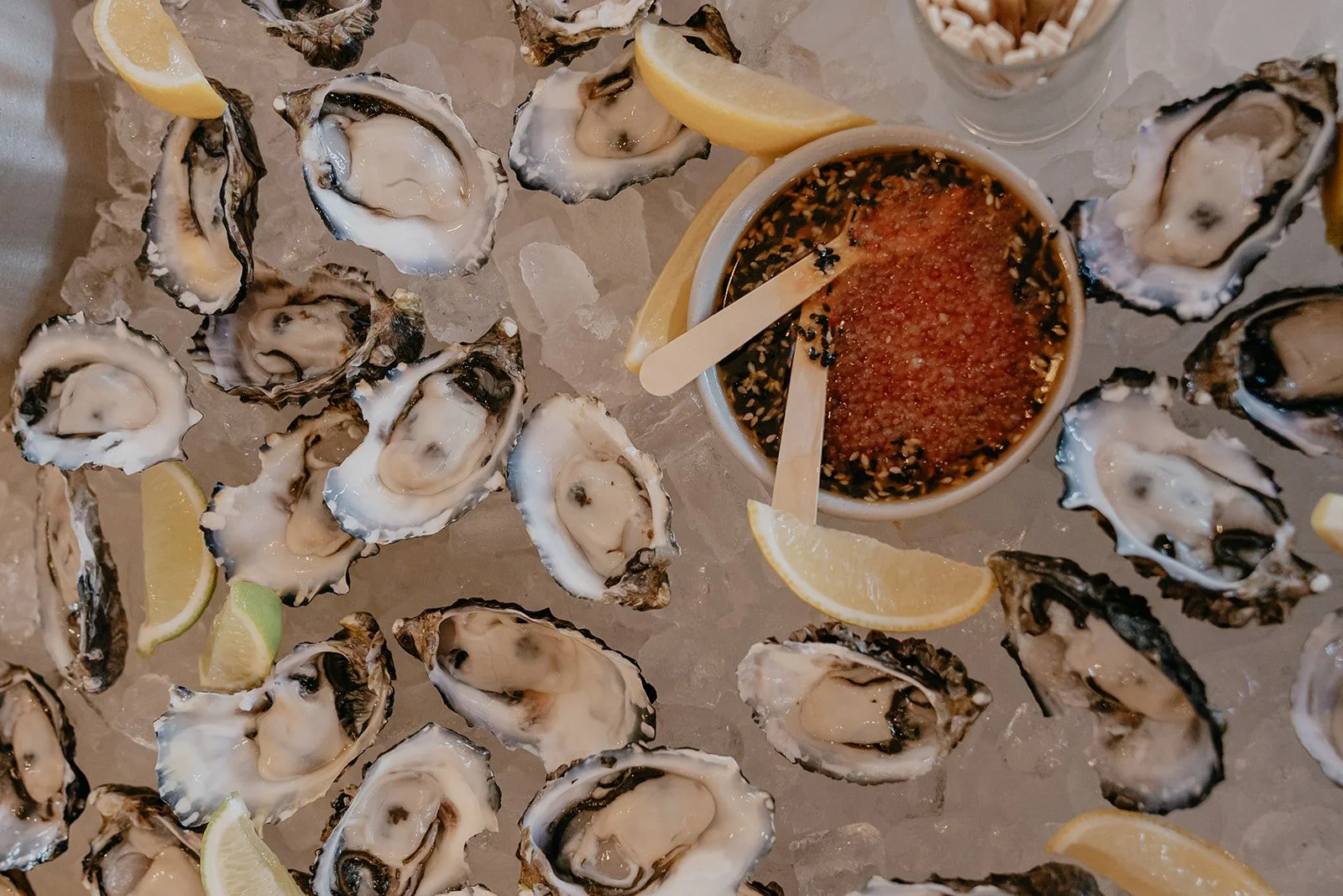 A platter of fresh oysters on ice with lemon wedges and a dipping sauce.
