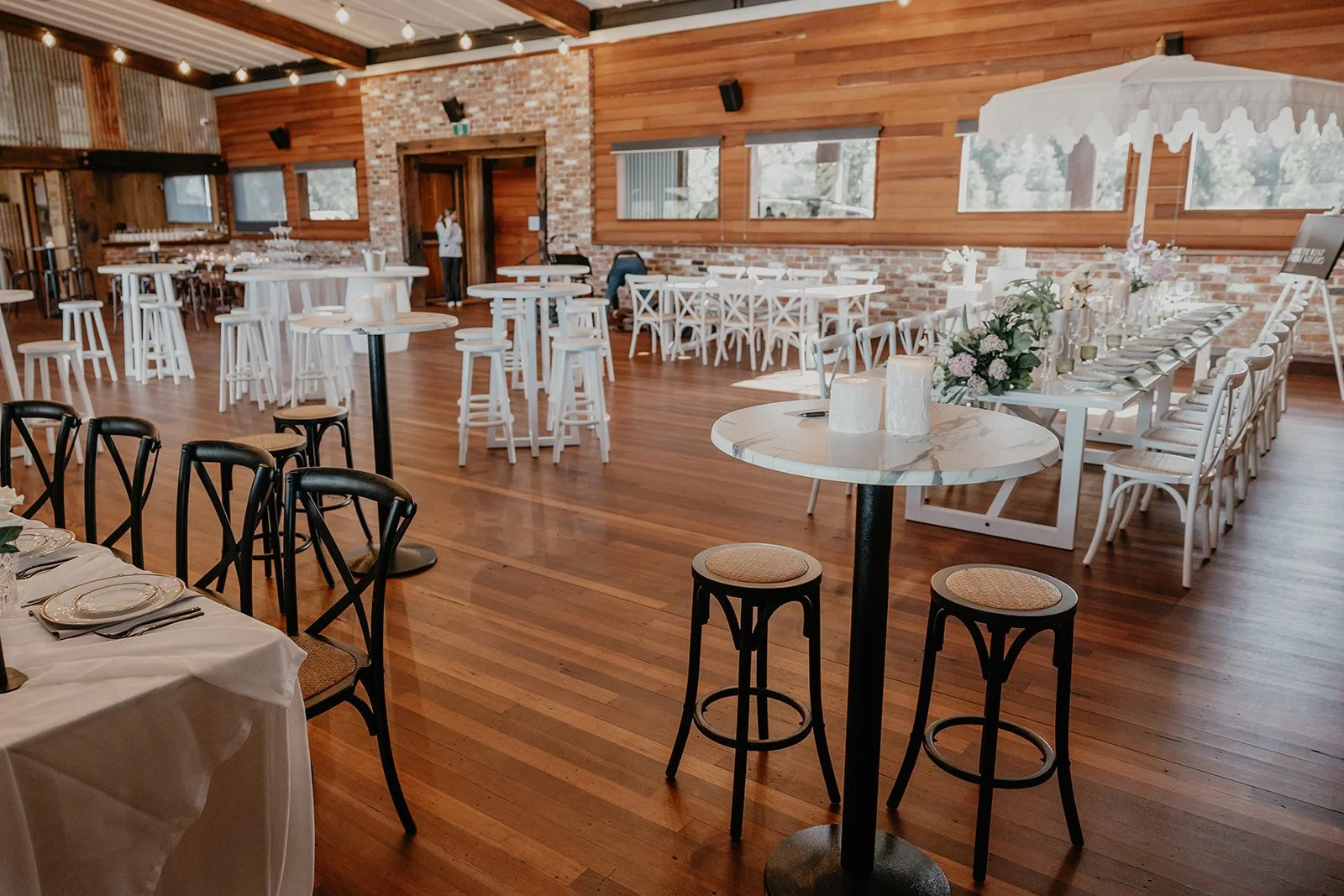 Empty wedding reception or event hall with wooden floors and walls, decorated with white tables, chairs, and floral centerpieces, some candles, and a large white umbrella over a table.