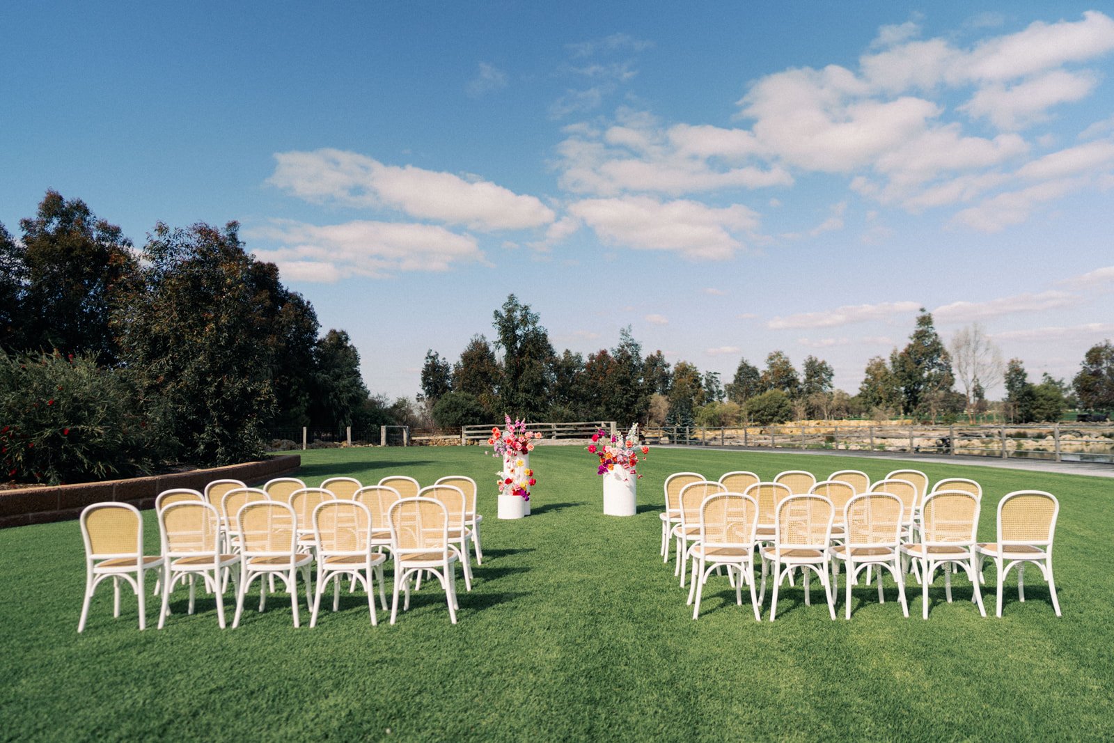 Outdoor wedding ceremony setup with rows of white chairs facing a floral arch on green grass, under a partly cloudy sky.