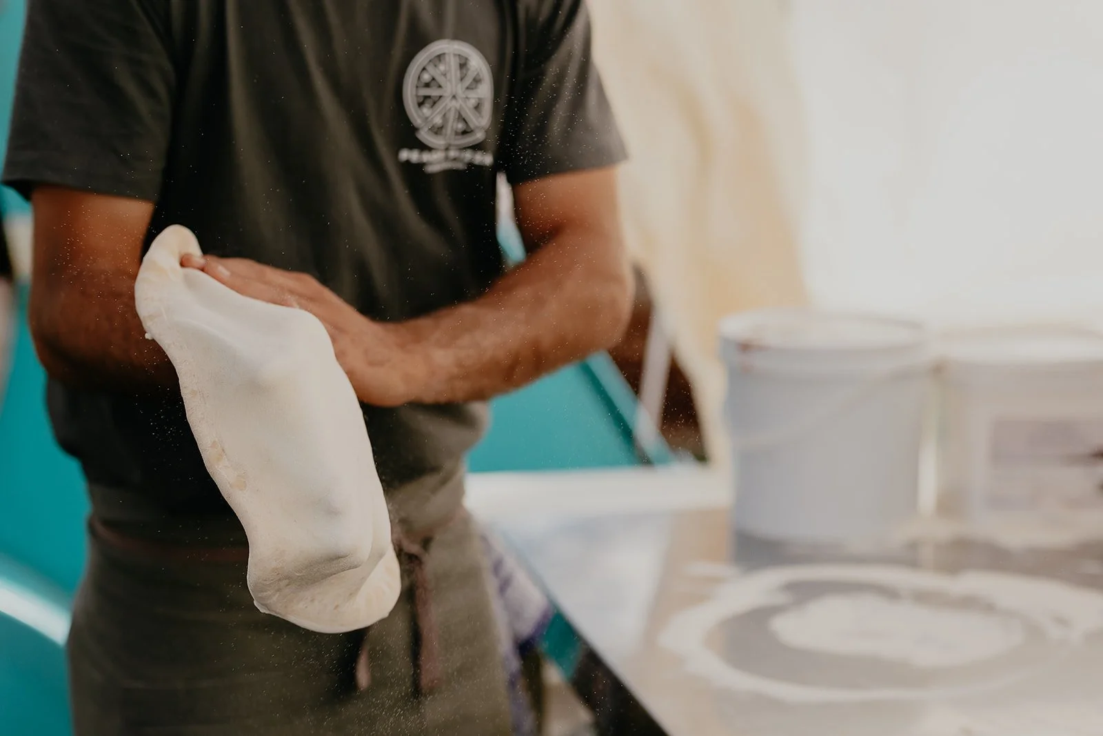 A person stretching and shaping pizza dough in a kitchen, with containers of ingredients nearby.