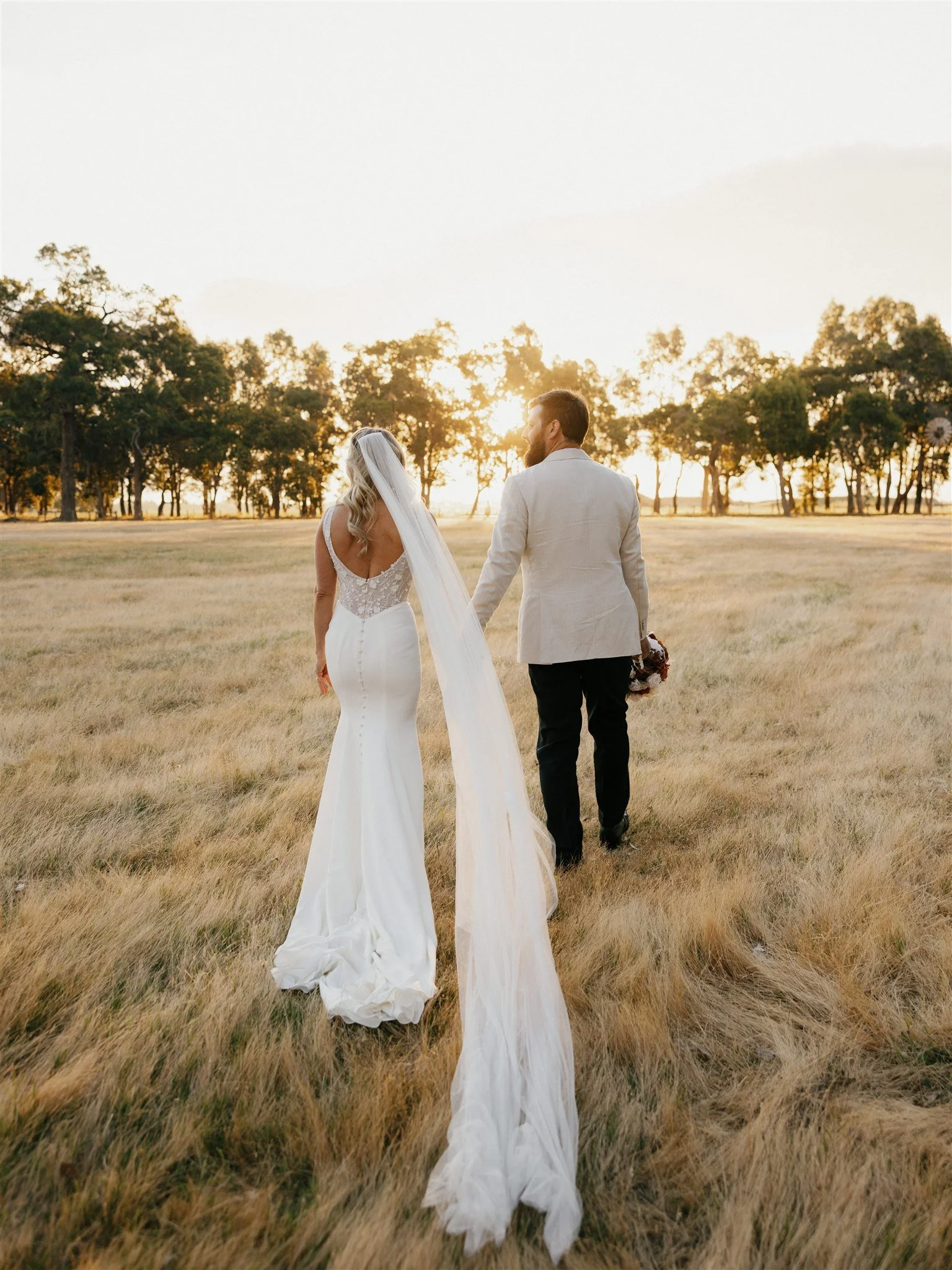 A bride and groom walking hand in hand through a grassy field at sunset, with trees in the background.
