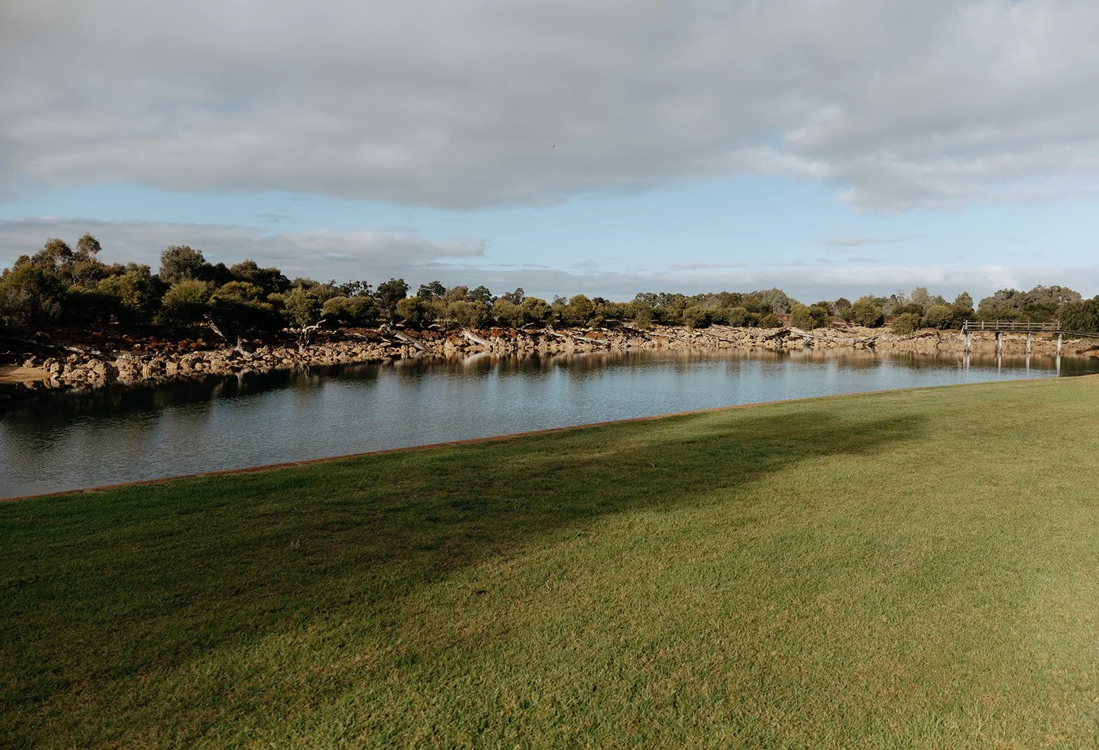 A grassy area next to a calm river with rocky banks, trees in the background, and partly cloudy sky.