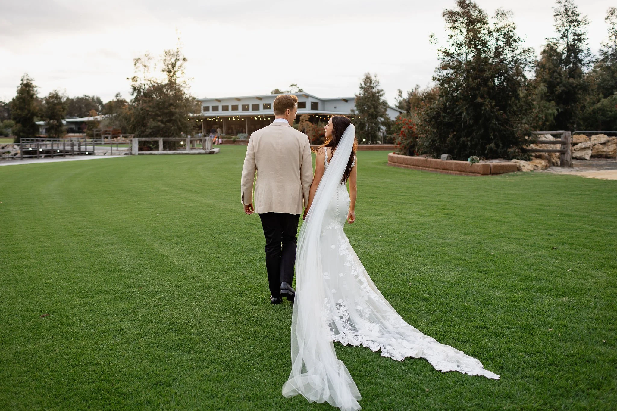 A bride and groom walking hand in hand on a lush green lawn during outdoor wedding, with a building and trees in the background.