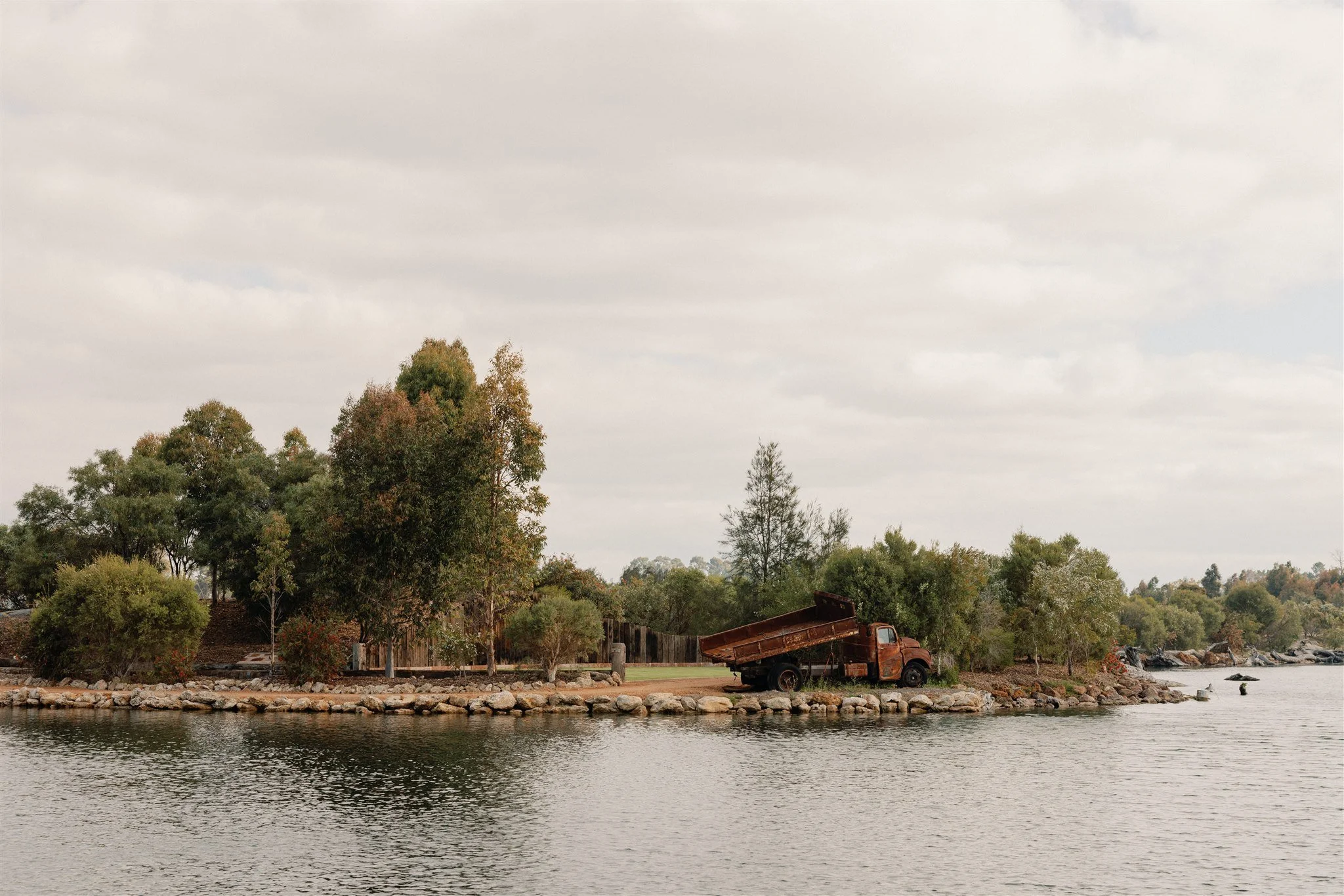 Old rusted Austin truck on the rocky shoreline of a lake on a rural property, with trees and a cloudy sky in the background.