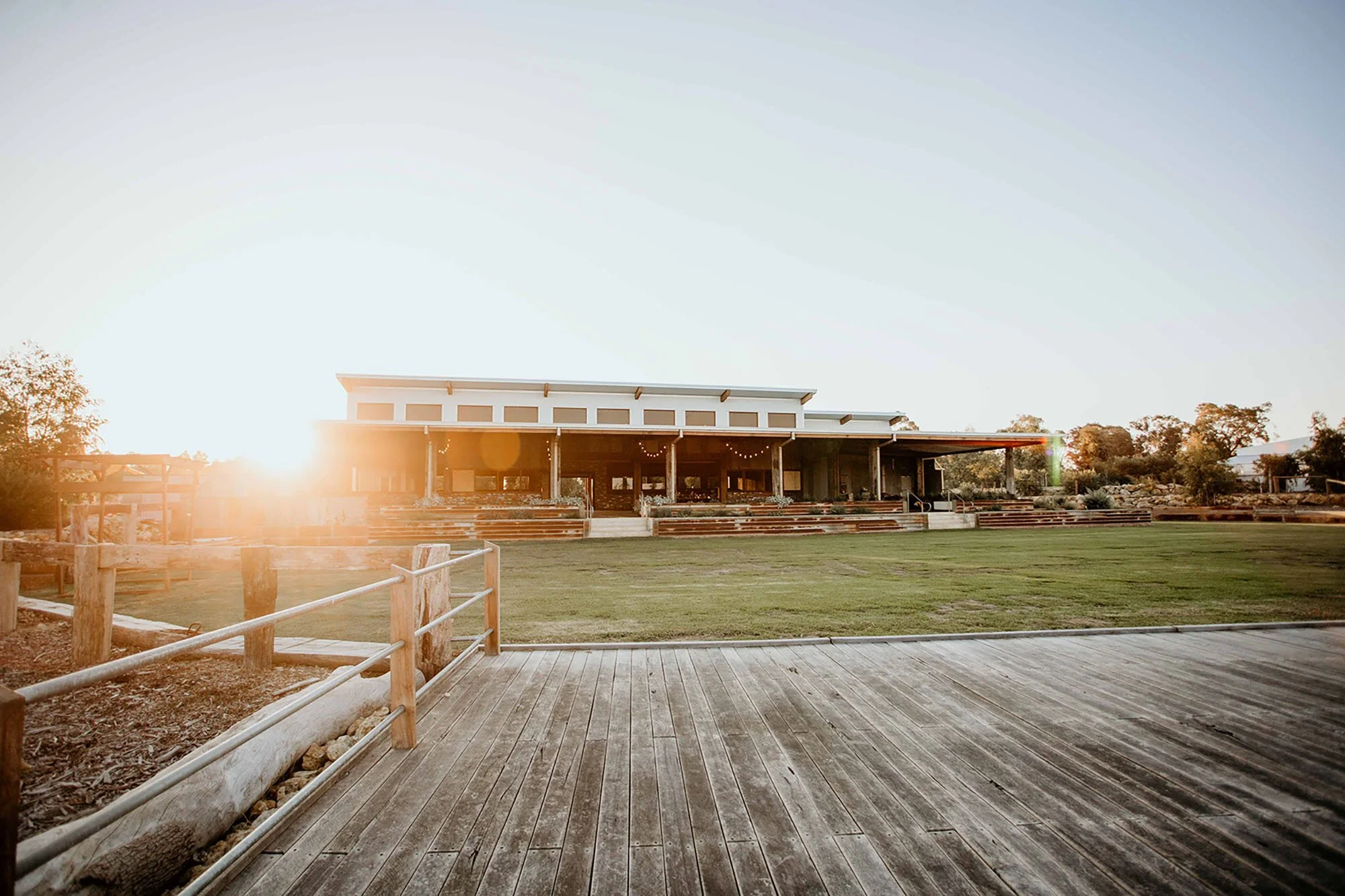 Sunset view of a modern building with a wide open veranda and tiered seating area, surrounded by a grassy field and wooden fence in the foreground.
