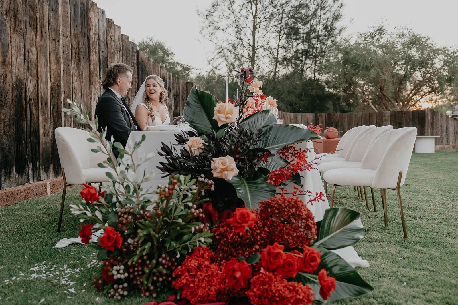 A wedding reception with a couple seated at a table outdoors. The table is decorated with a large flower arrangement featuring red, peach, and dark foliage. The background shows a rustic wooden fence, grassy yard, and trees at sunset.